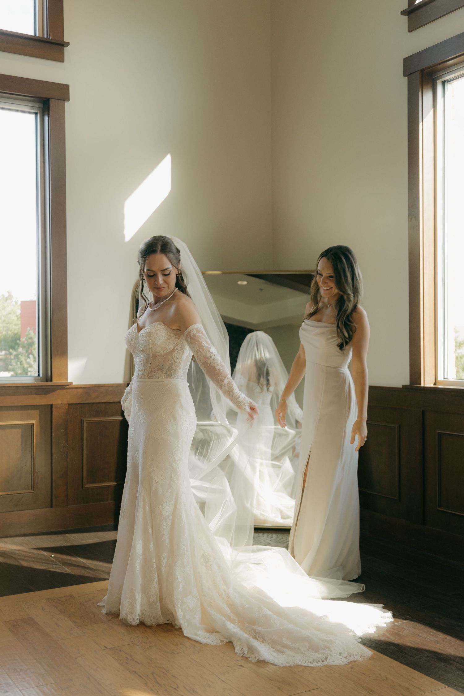 Bride in lace wedding gown with veil being helped by bridesmaid in elegant sunlit room.