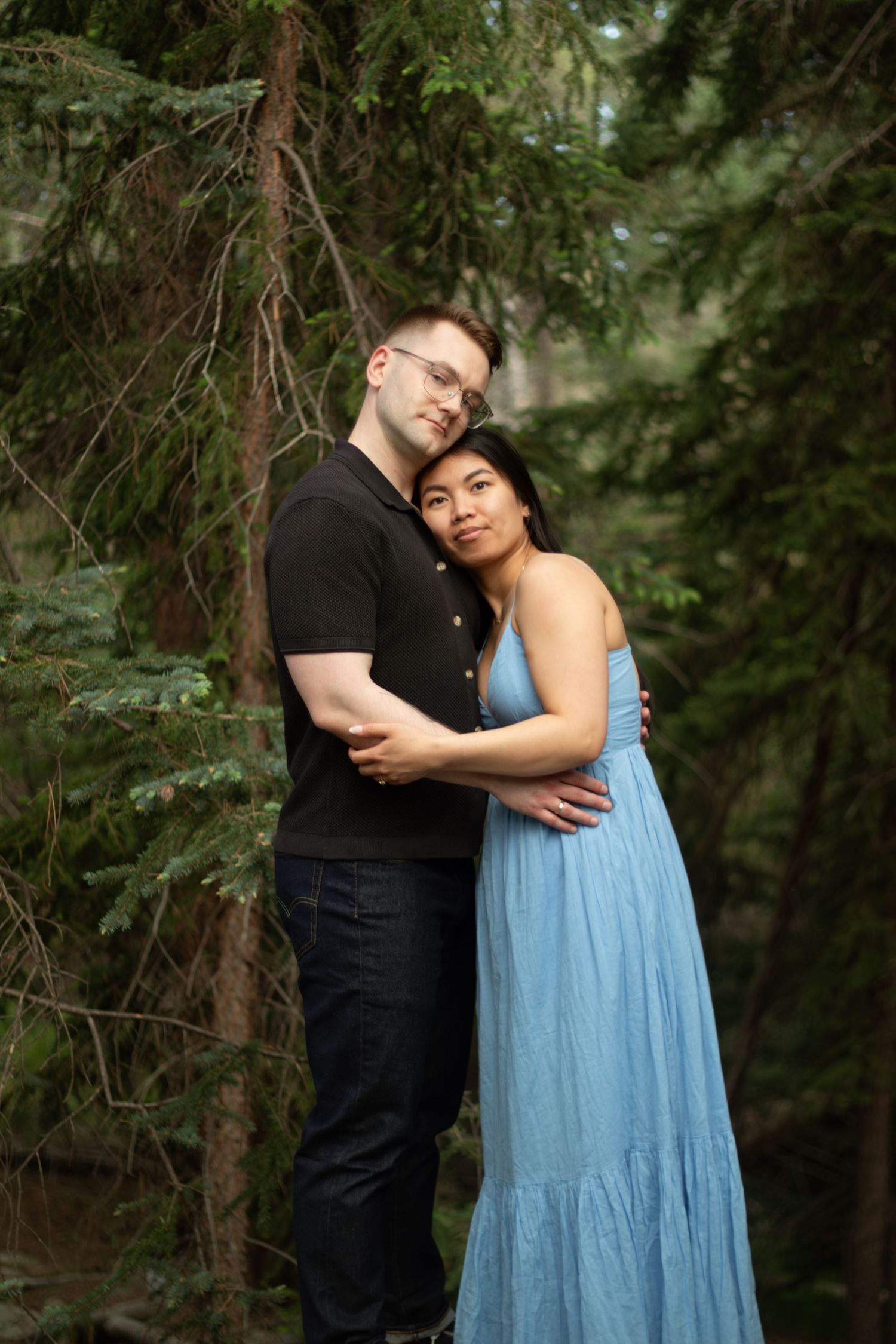 Two people share an intimate moment while standing among pine trees, dressed in black and light blue.