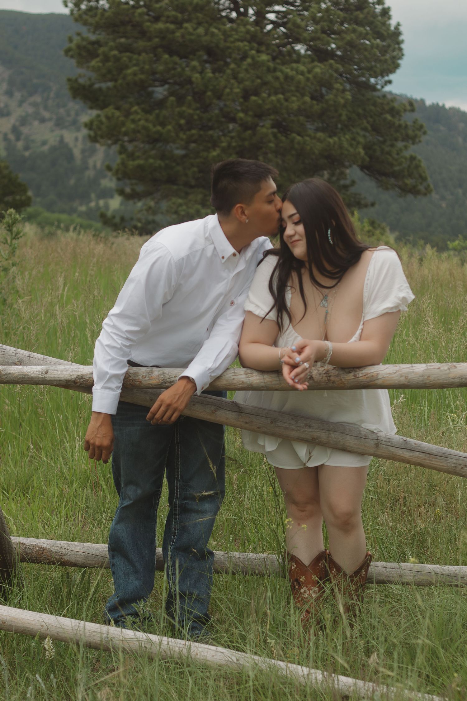 Couple in white clothing embracing near wooden fence rails with mountains and pine trees in background.