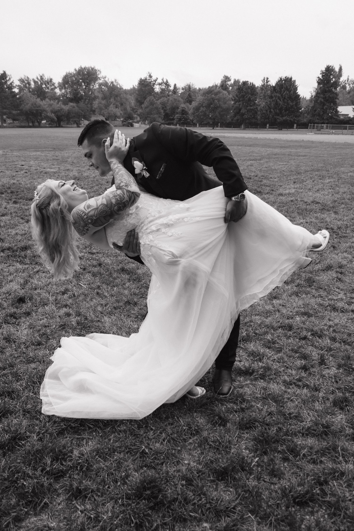 Two people in formal attire perform a romantic dip pose on a grassy field in black and white.