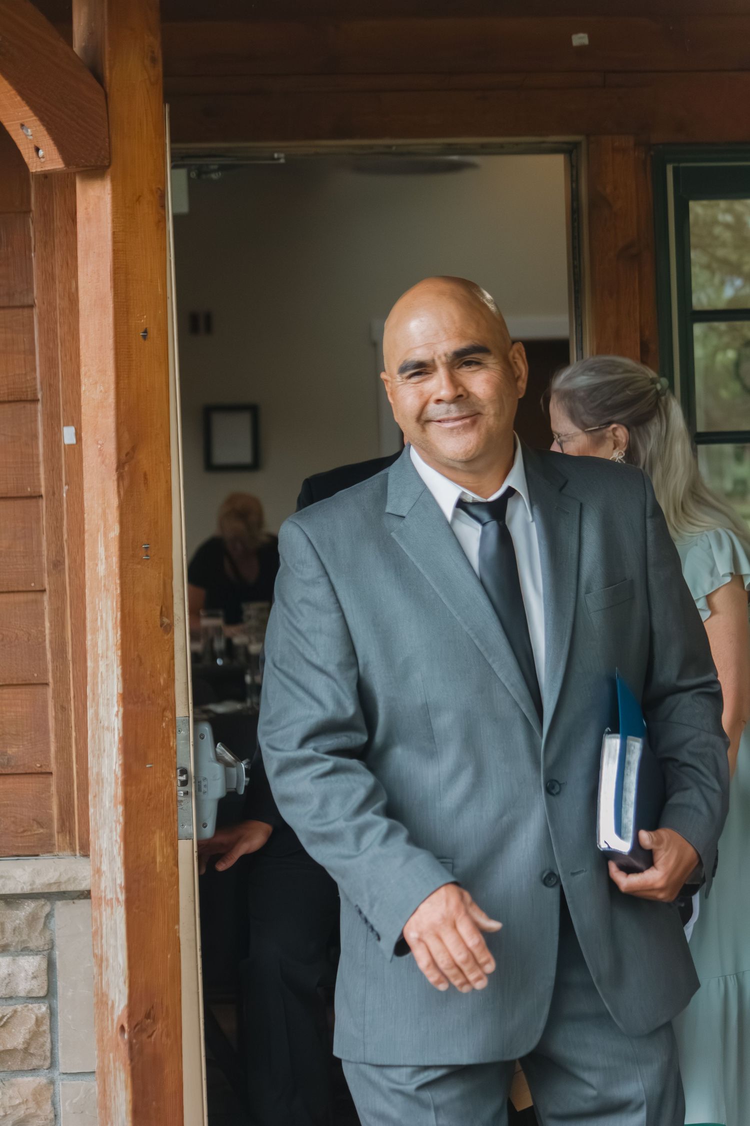 A wedding guest in a gray suit smiles warmly while standing in a doorway.