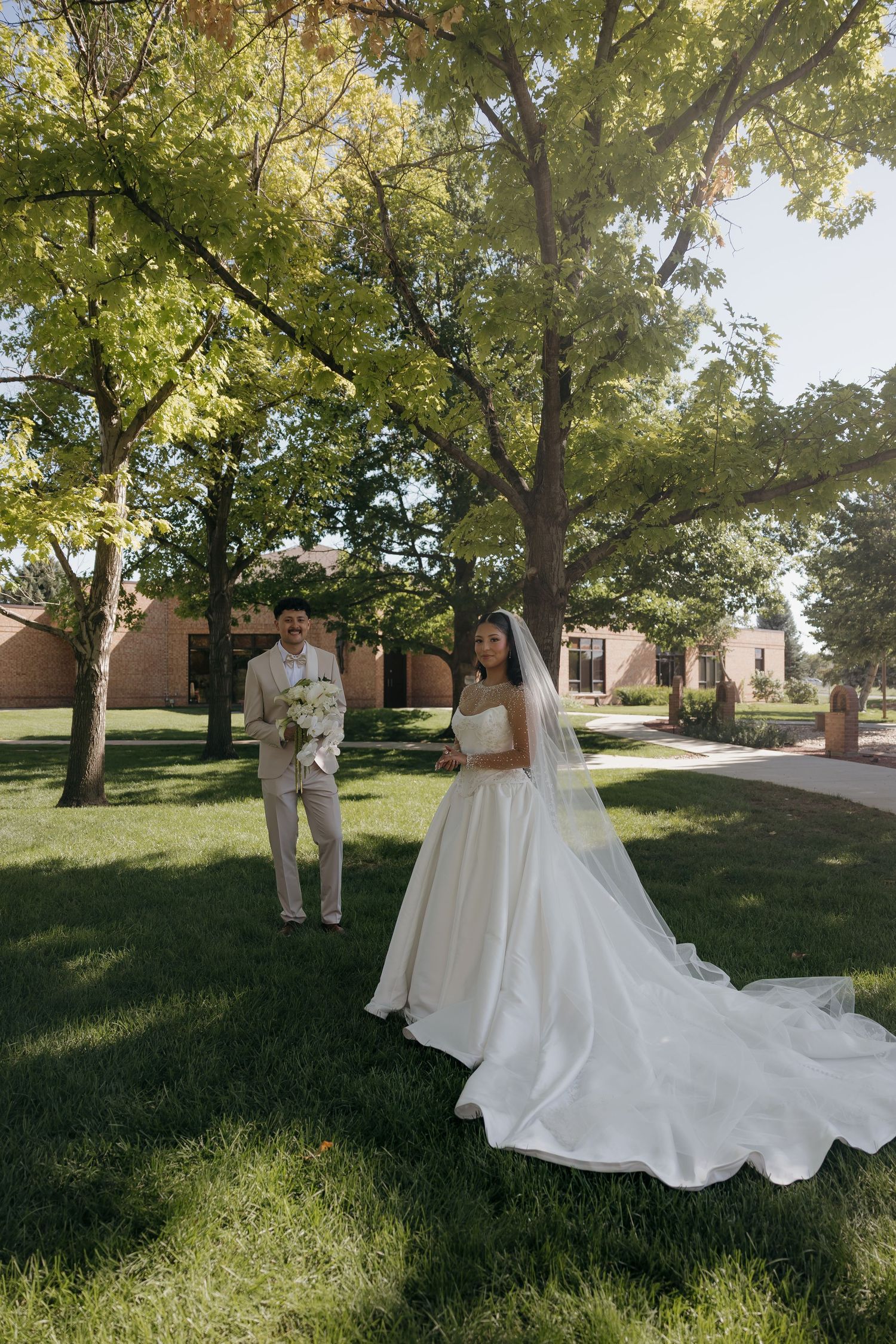 Bride in flowing white gown with veil and groom in tan suit stand on sunlit grass beneath green trees outside brick church.