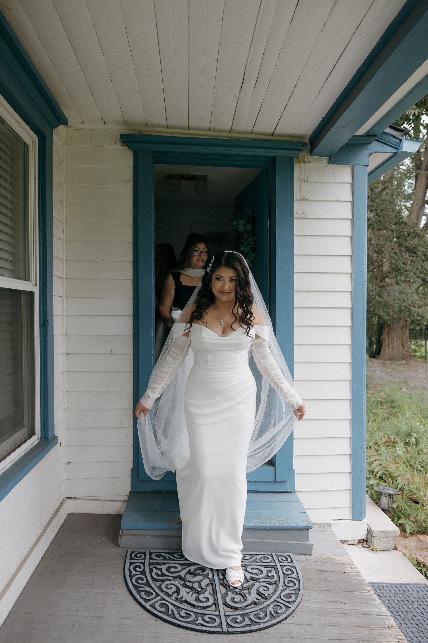 Bride in white fitted gown with lace sleeves and veil steps onto porch of white house with blue trim.