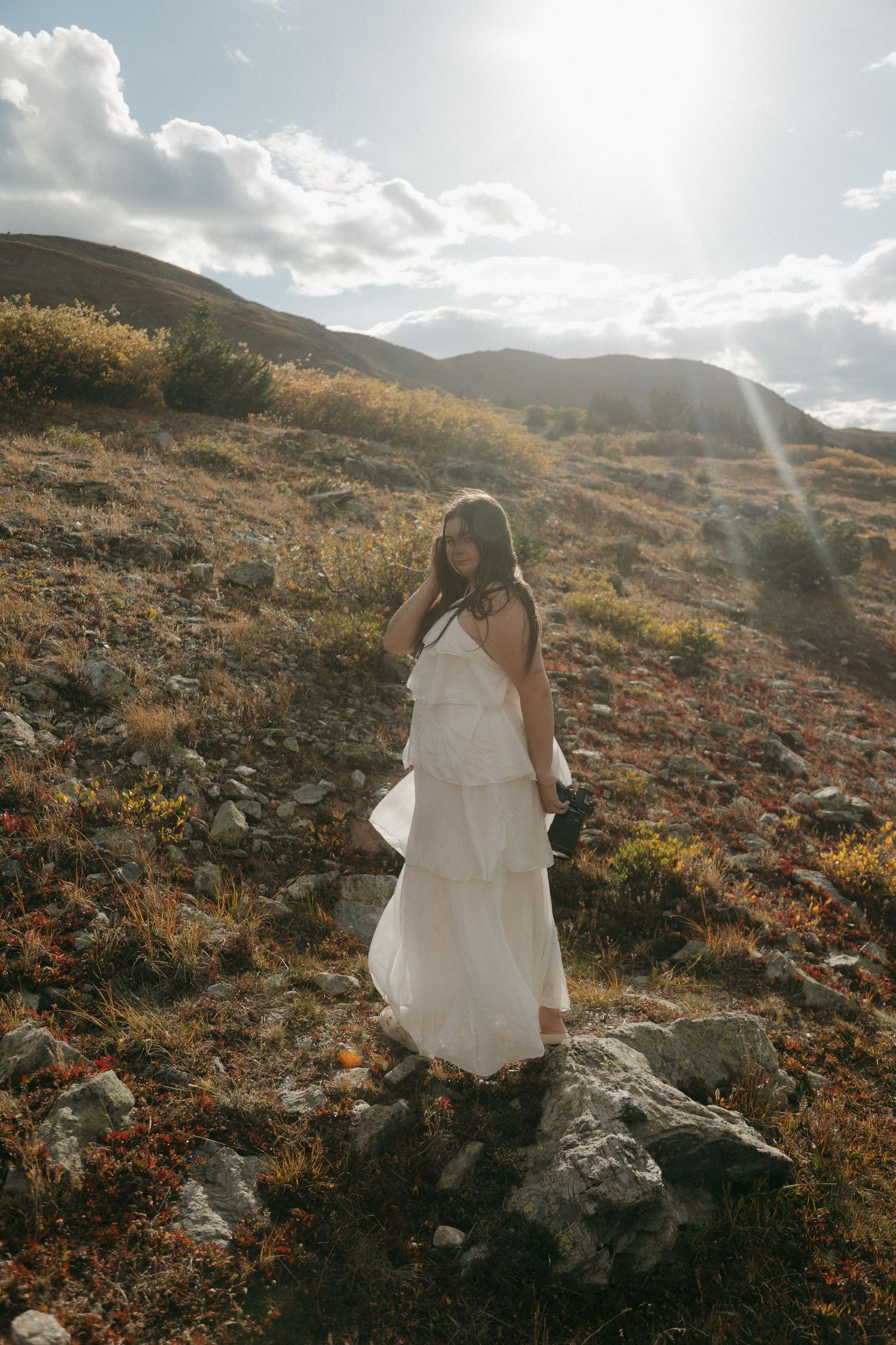 Woman in white tiered dress standing on rocky mountain hillside with golden grass and sun rays in background.