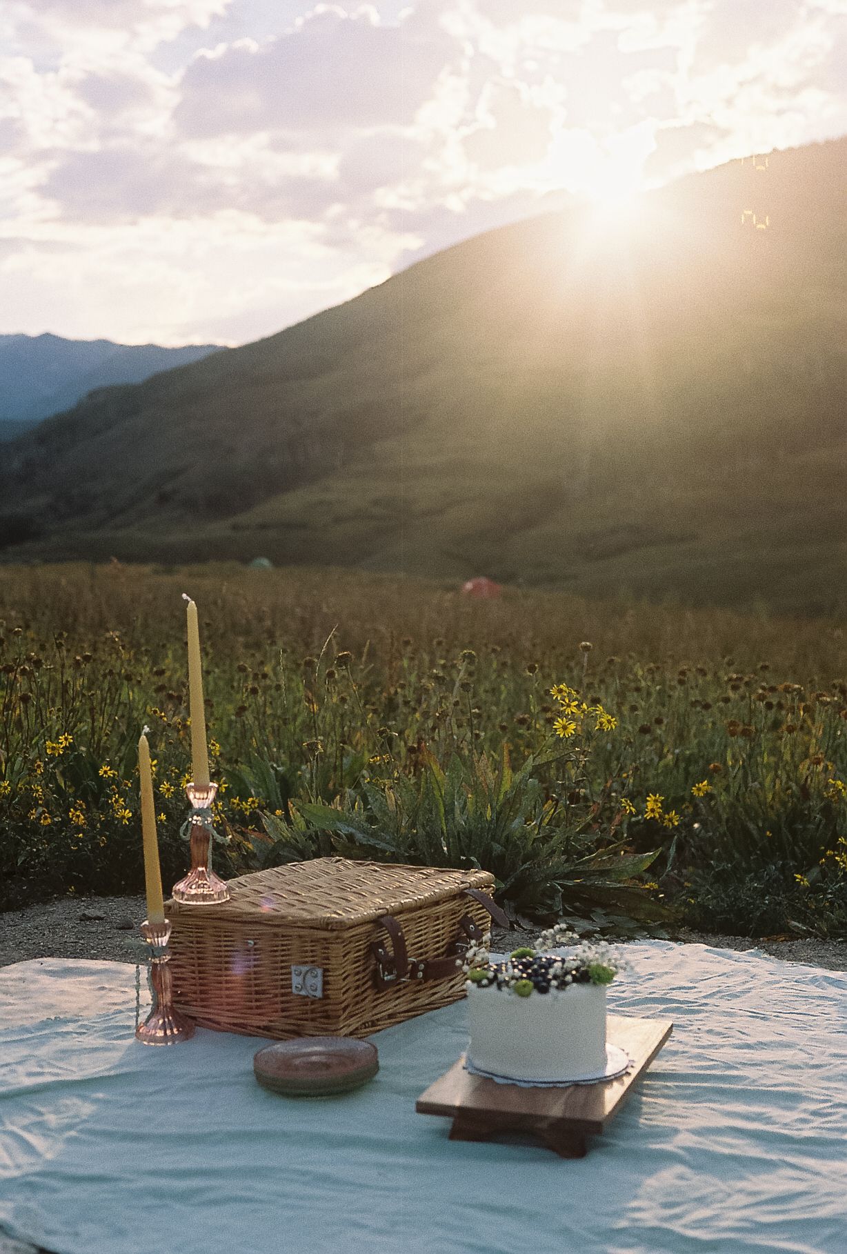Rustic wedding picnic setup with cake, wicker basket, and candle in a sunflower field at golden sunset.