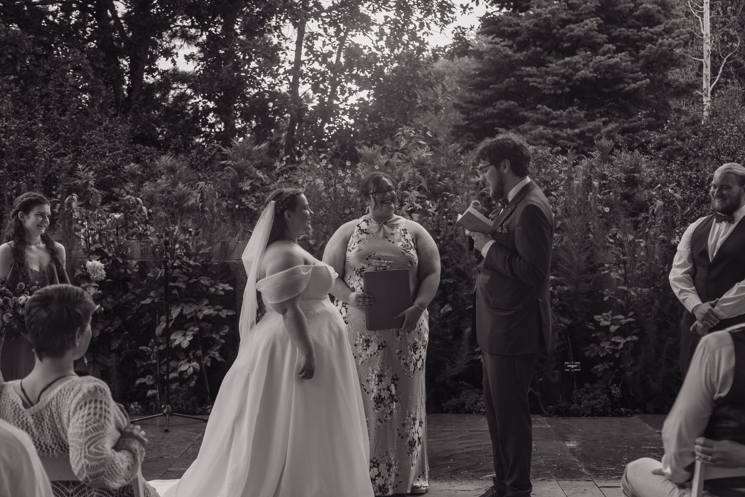 Black and white outdoor wedding ceremony with bride in white gown, groom, and officiant surrounded by seated guests and lush trees.
