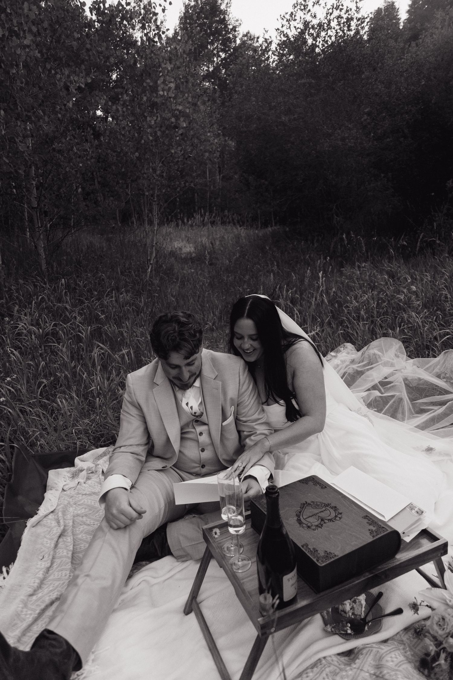 Black and white photo of bride and groom reading letters together on blanket during intimate outdoor forest wedding picnic.