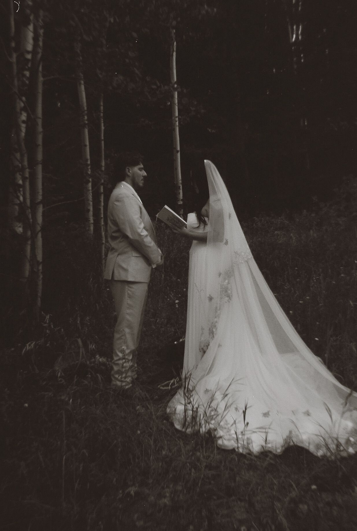 Black and white photo of bride and groom exchanging vows among birch trees in a dark forest.