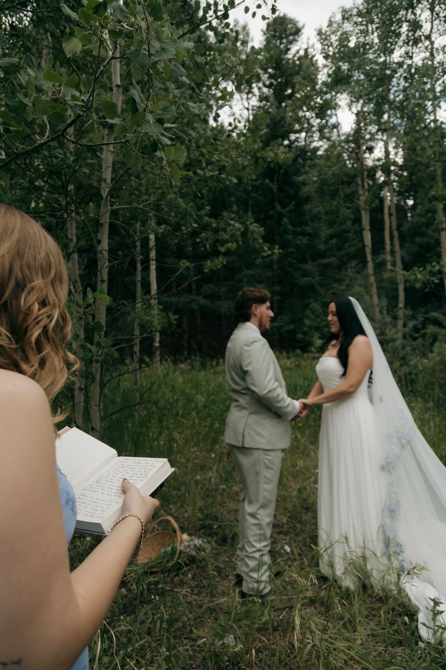Couple exchanges vows in intimate outdoor forest wedding ceremony as officiant reads from book.