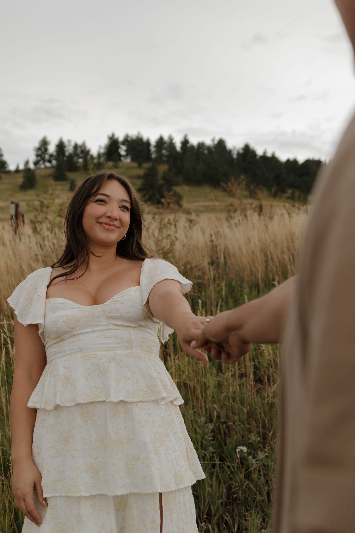 Woman in white floral tiered dress holding mans hand in lush green meadow, smiling warmly.