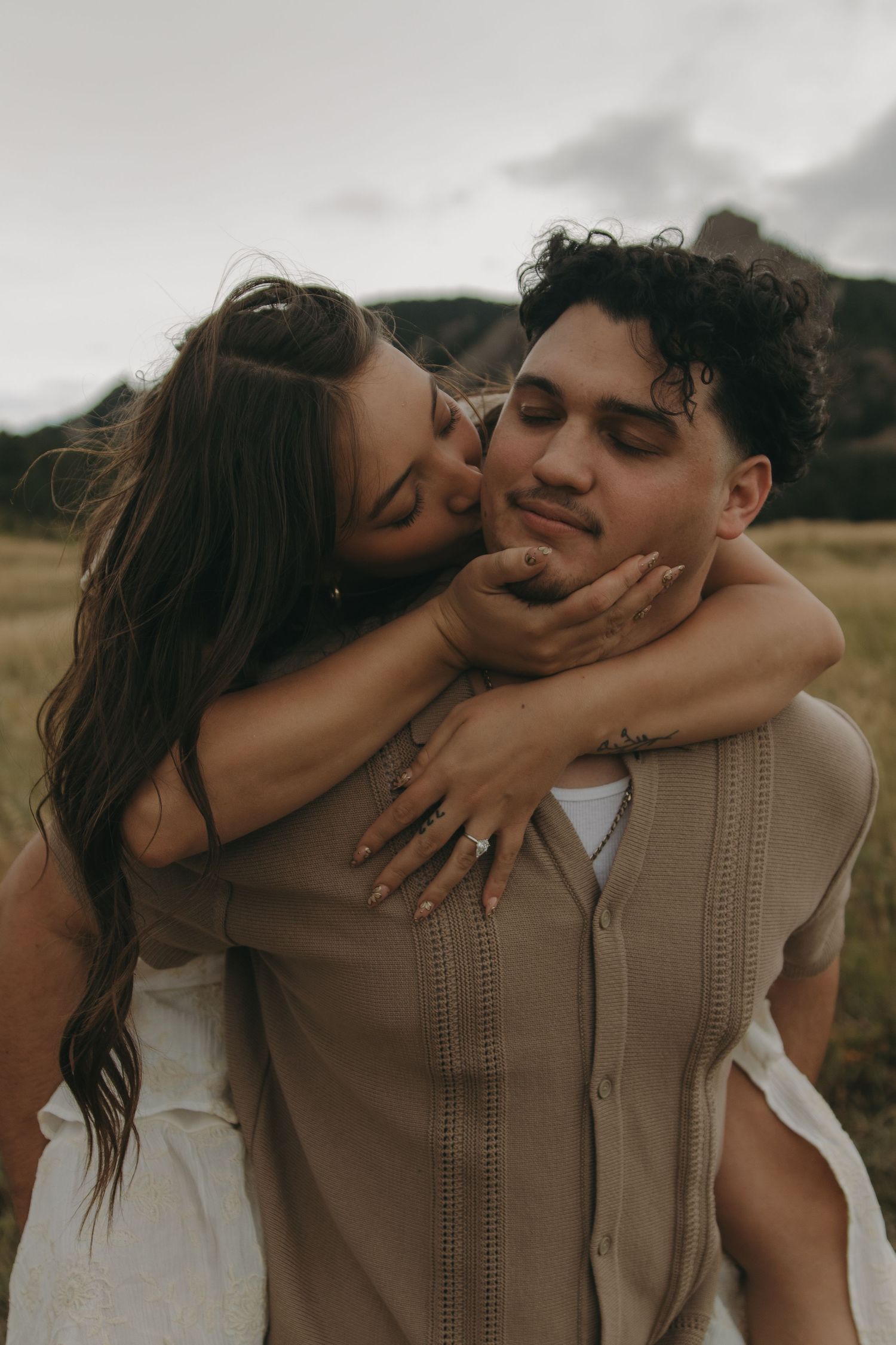 A woman hugs a man from behind and kisses his cheek in a scenic outdoor field with mountains.