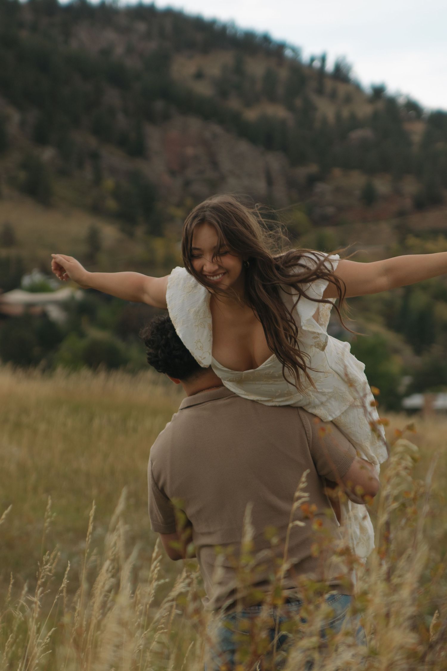 A smiling woman spreads her arms wide while being carried on a man's shoulders in a mountain meadow.