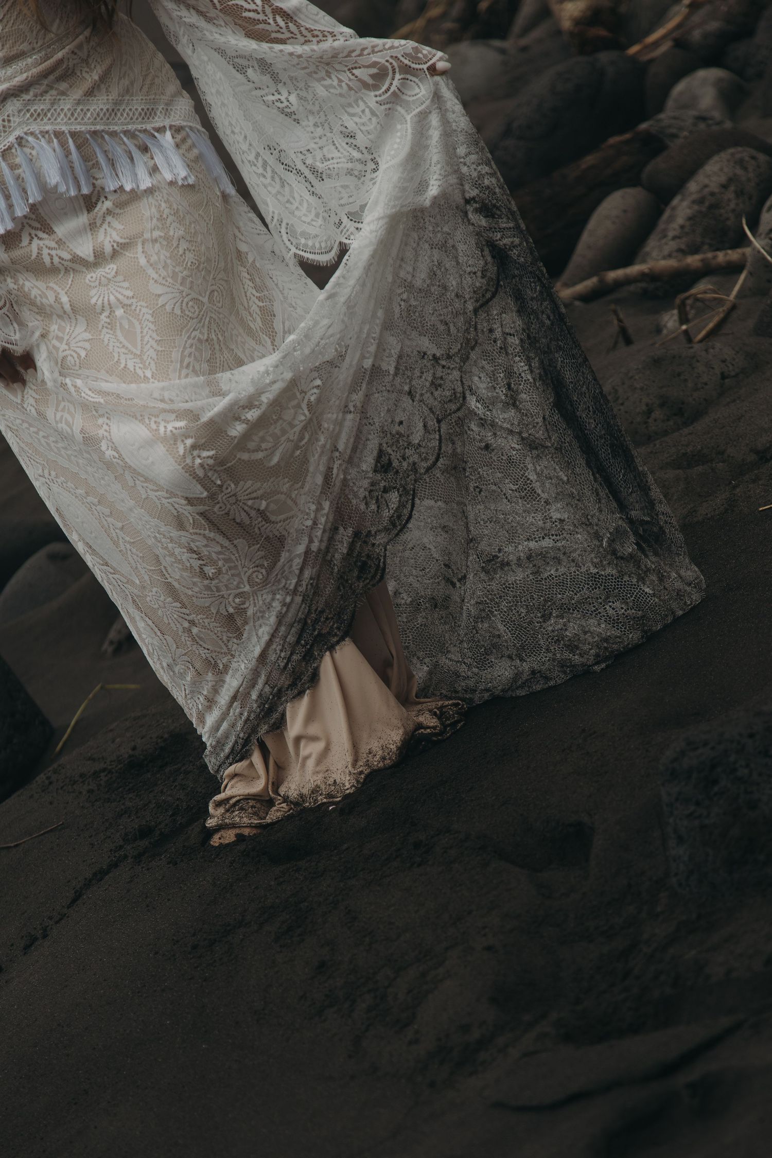 Close-up detail of a wet bohemian lace wedding dress hem on dark sand beach with rocks nearby.