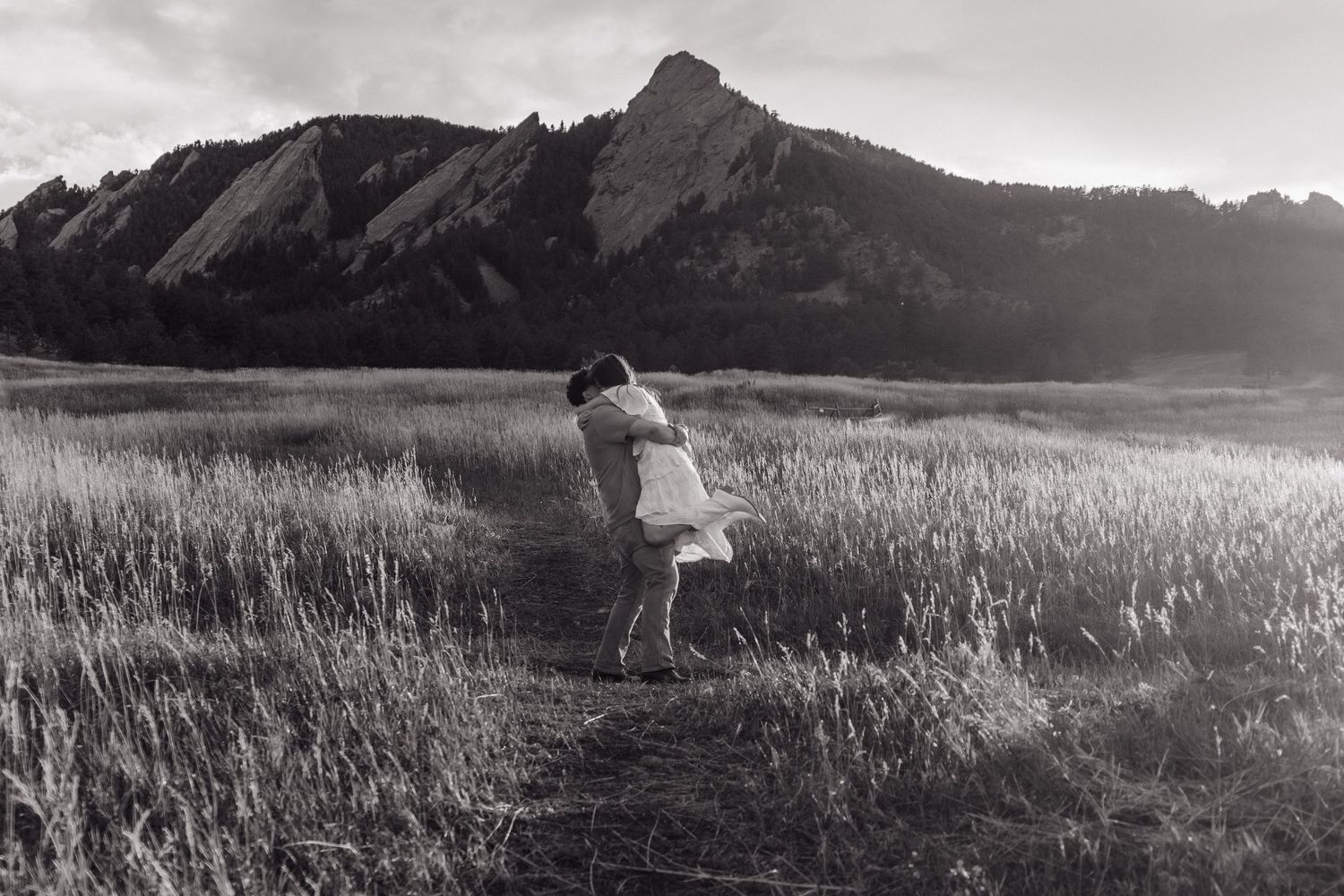 Black and white photo of couple embracing in tall grass field with dramatic Flatirons mountain backdrop.