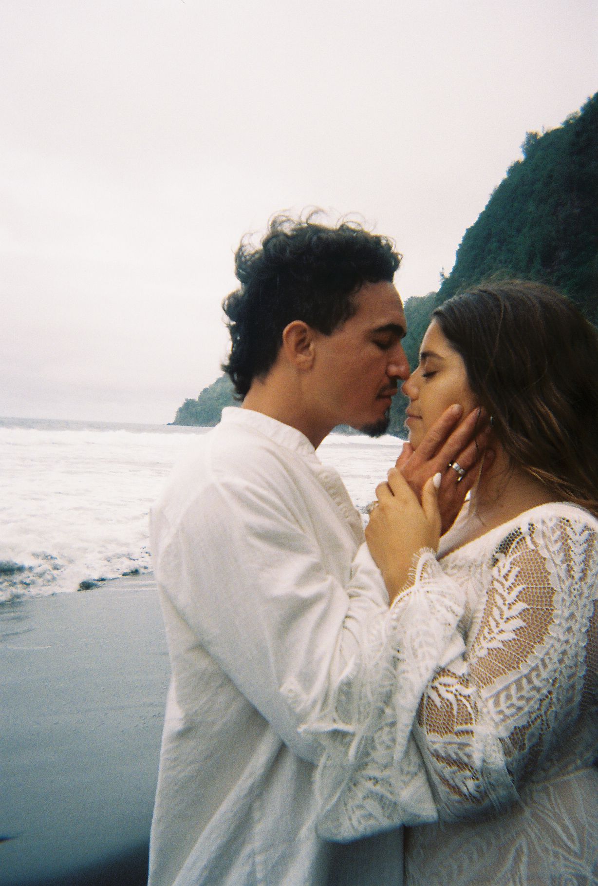 Couple sharing romantic kiss on black sand beach with dramatic green sea cliffs behind them.