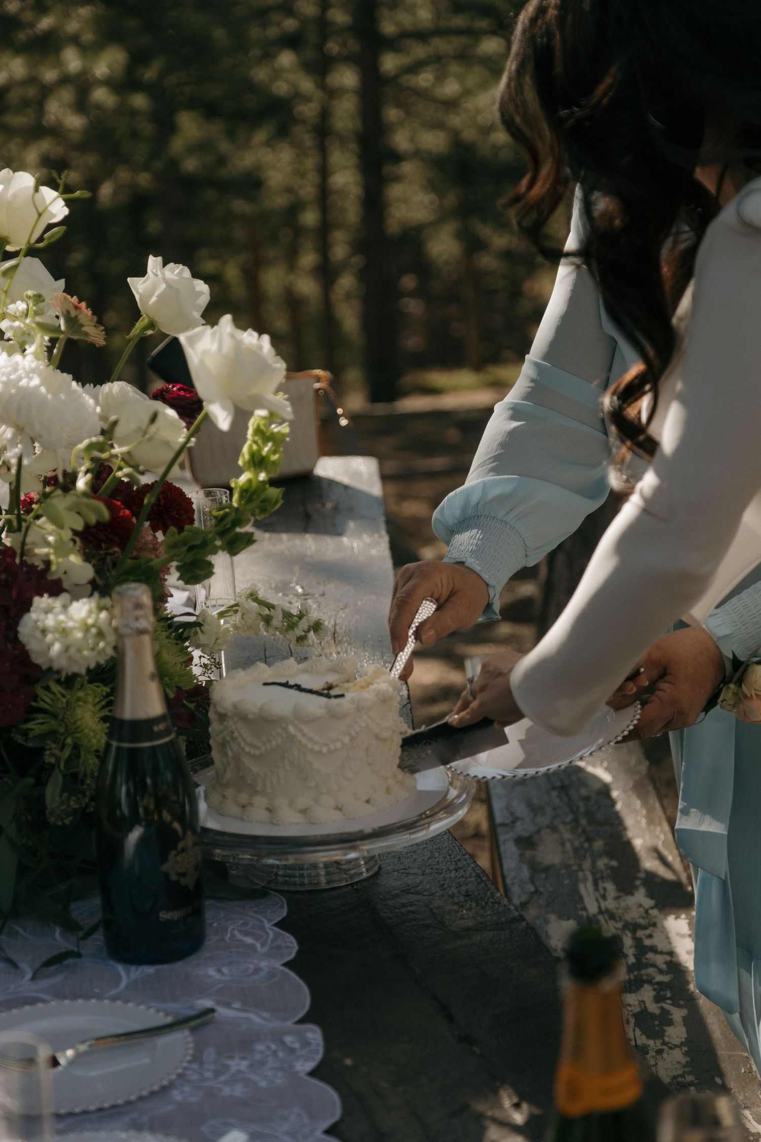 Bride in white dress cuts wedding cake on outdoor table with champagne and flowers in forest setting.