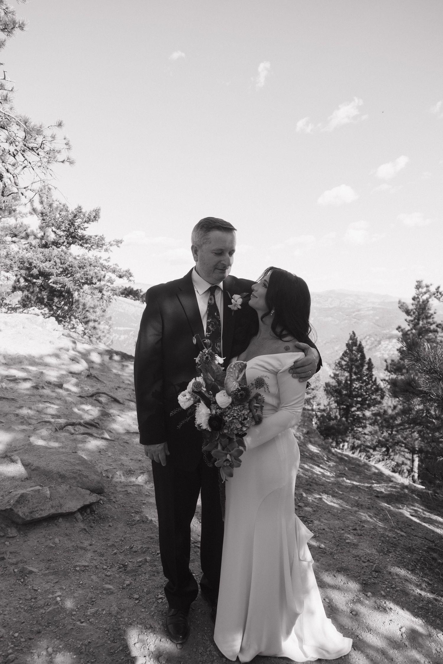 Black and white photo of bride and groom smiling together on mountain overlook with trees.