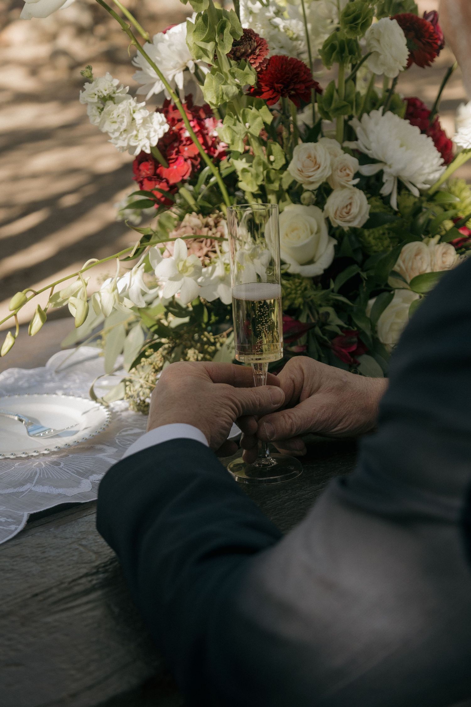 Groom's hands hold a champagne flute beside a lush floral arrangement of red and white roses at a wedding table.