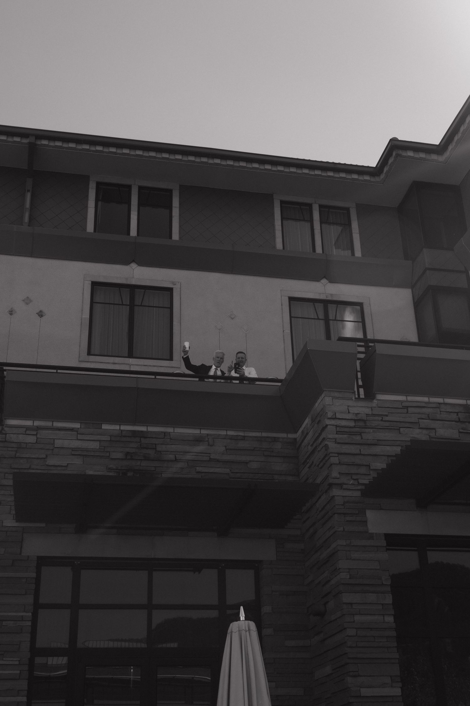 Black and white photo of two people waving from hotel balcony above stone facade.