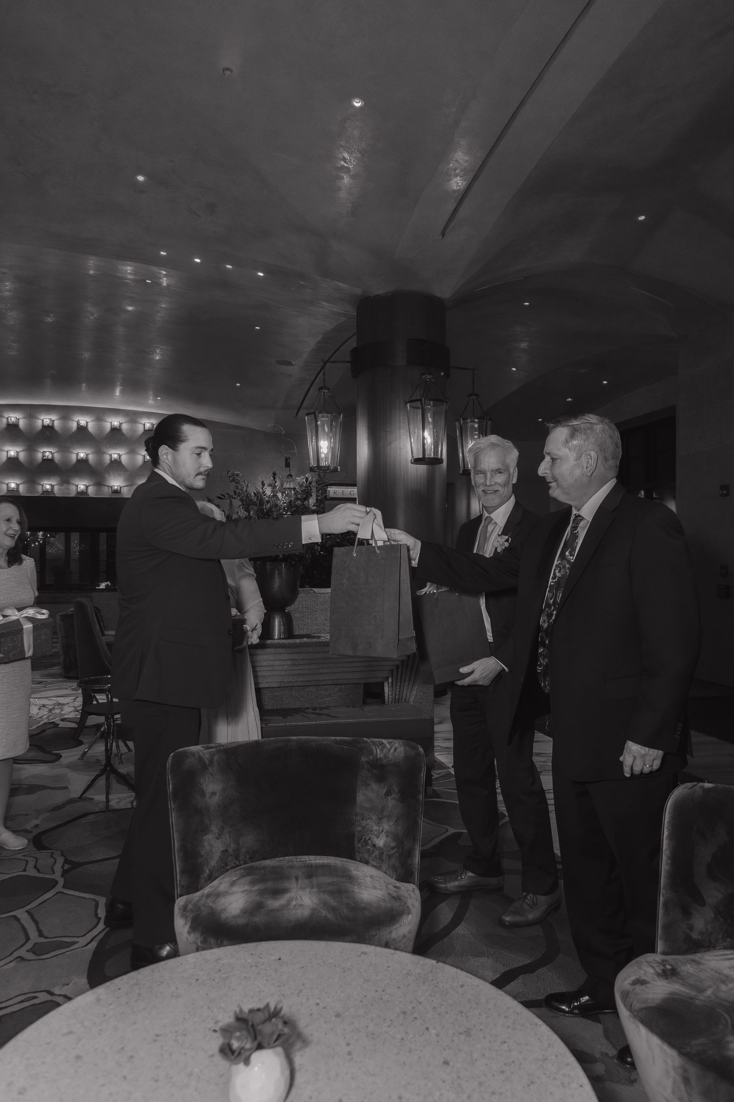 Three men in suits exchange gift bags in an elegant dimly lit bar or lounge setting.