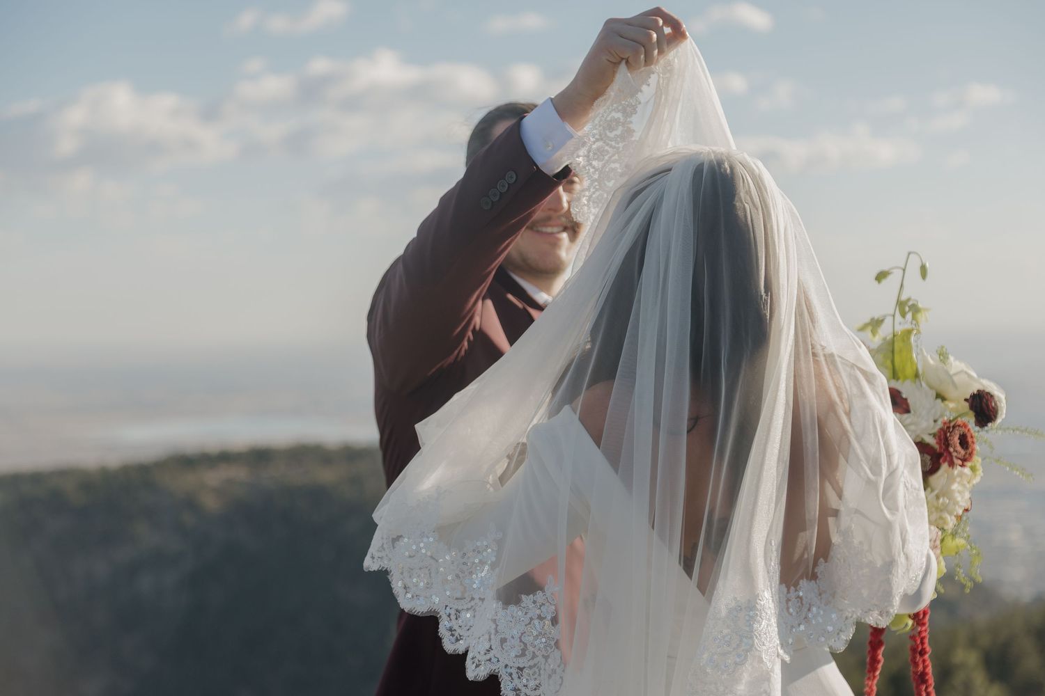 A groom lifts the lace-edged bridal veil over the couple as they embrace with scenic views behind them.