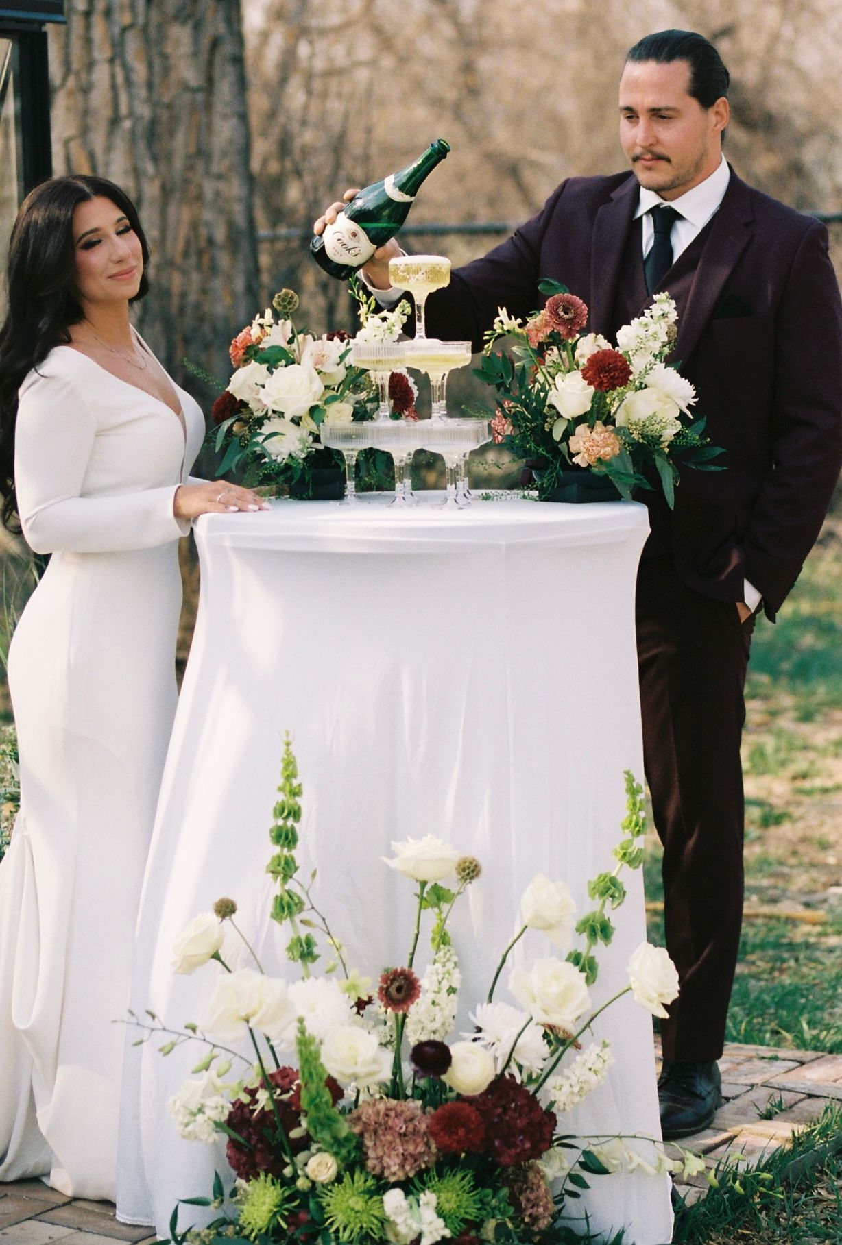 Bride and groom pouring champagne tower at outdoor wedding reception with white and burgundy florals.