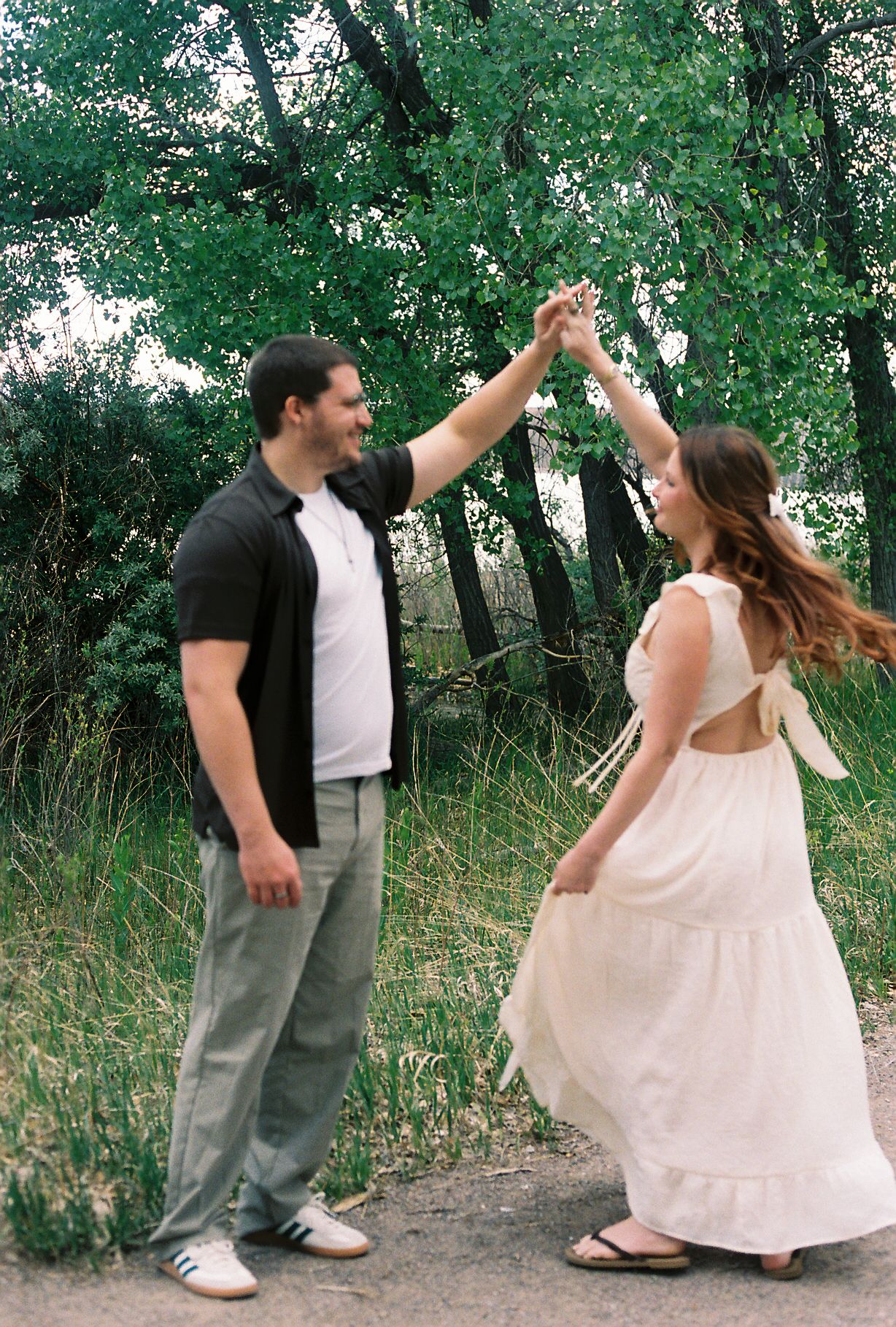 Smiling couple dancing together on a path surrounded by green trees in natural light.