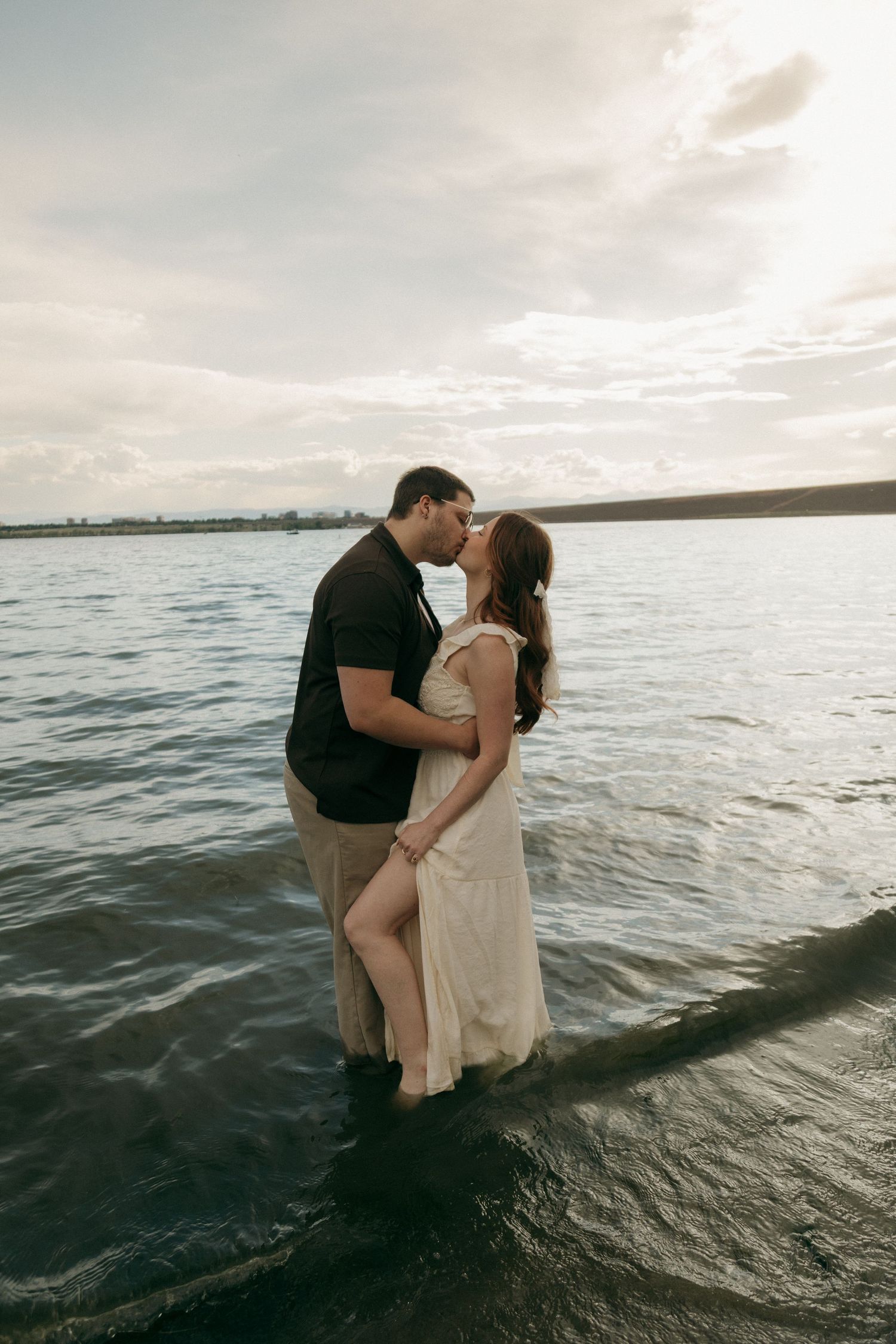 Romantic couple sharing a kiss while standing in a lake at golden hour with cloudy skies behind them.