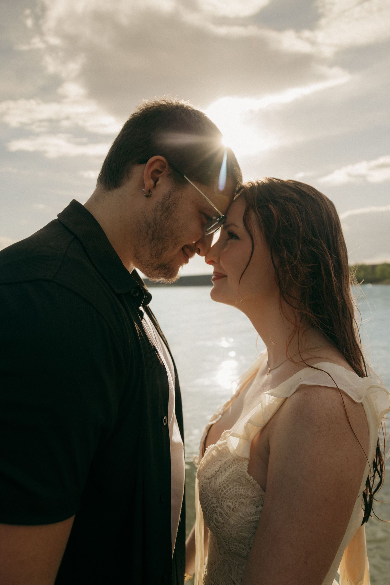 Romantic couple touching foreheads near a sunlit lake, man in black shirt and woman in white lace dress, golden backlight.