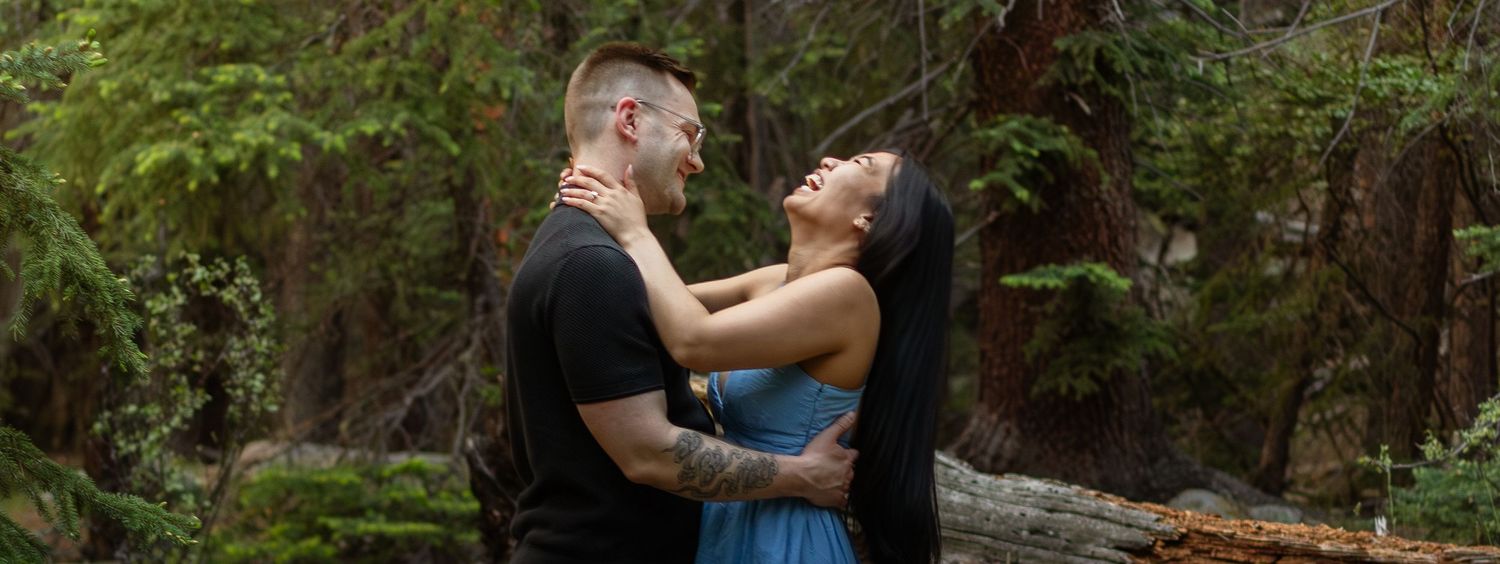 A couple laughs together during a romantic moment in a wooded mountain setting.