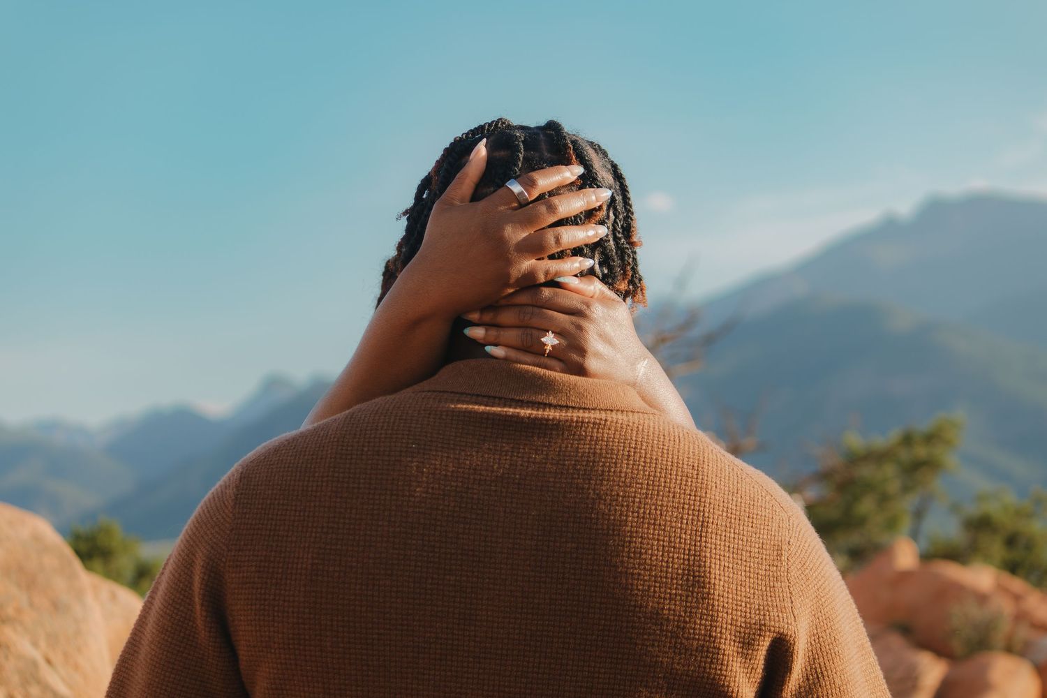 Person wearing brown sweater stands against scenic mountain backdrop with blue skies during autumn season.