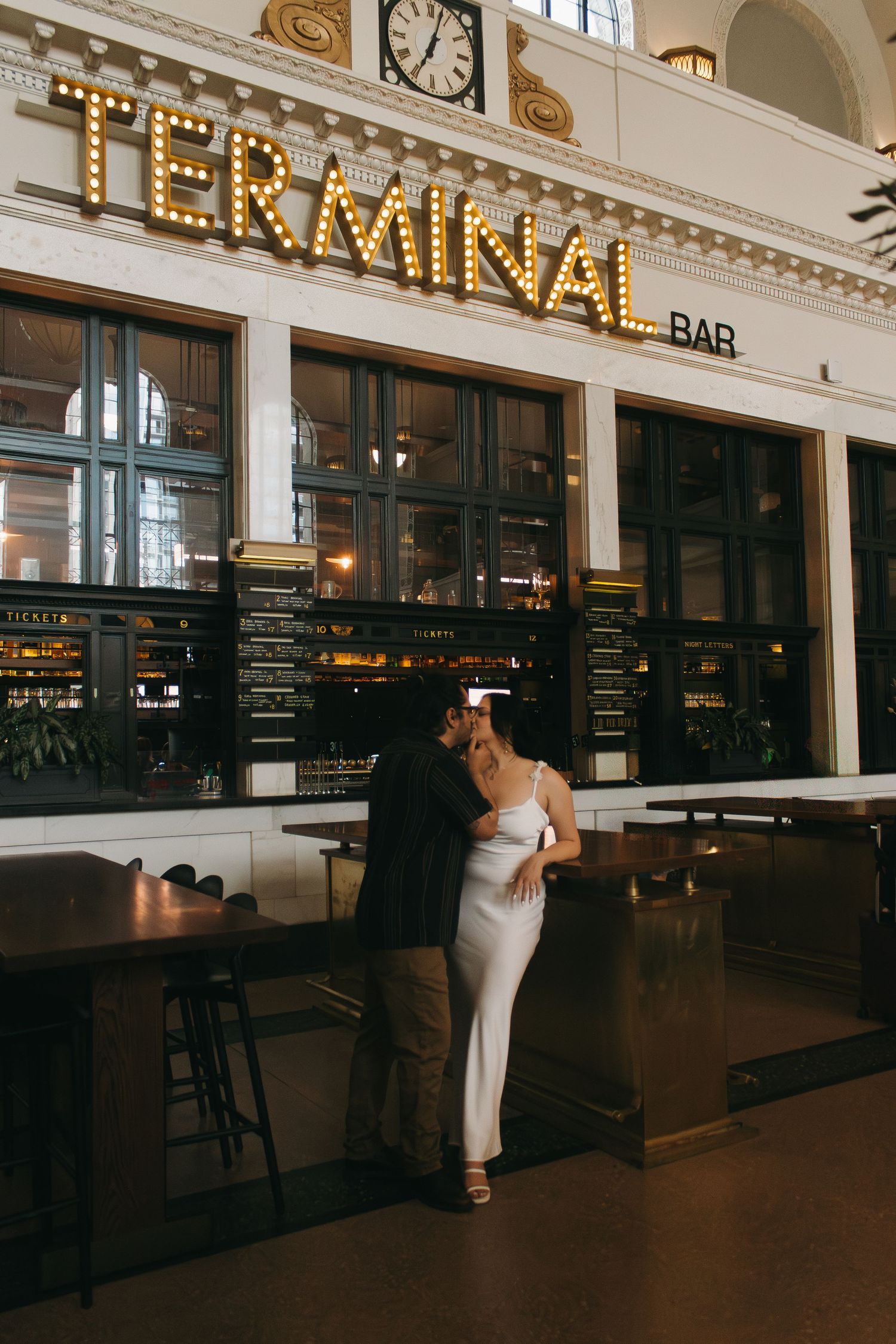 A couple embraces at the Terminal Bar, with illuminated vintage signage and classic architecture in the background.