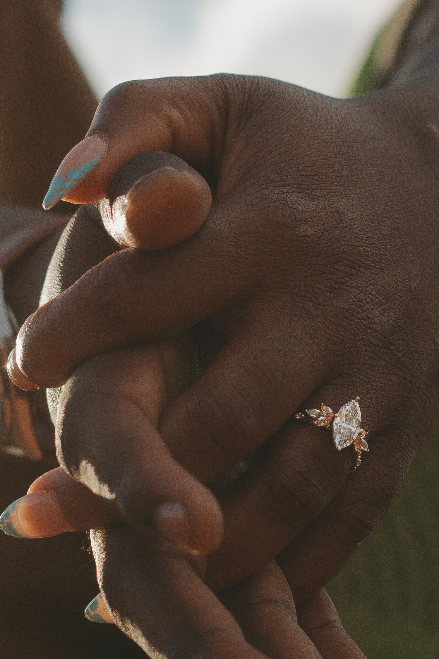 Close-up of intertwined hands showing a sparkling engagement ring in warm sunlight.