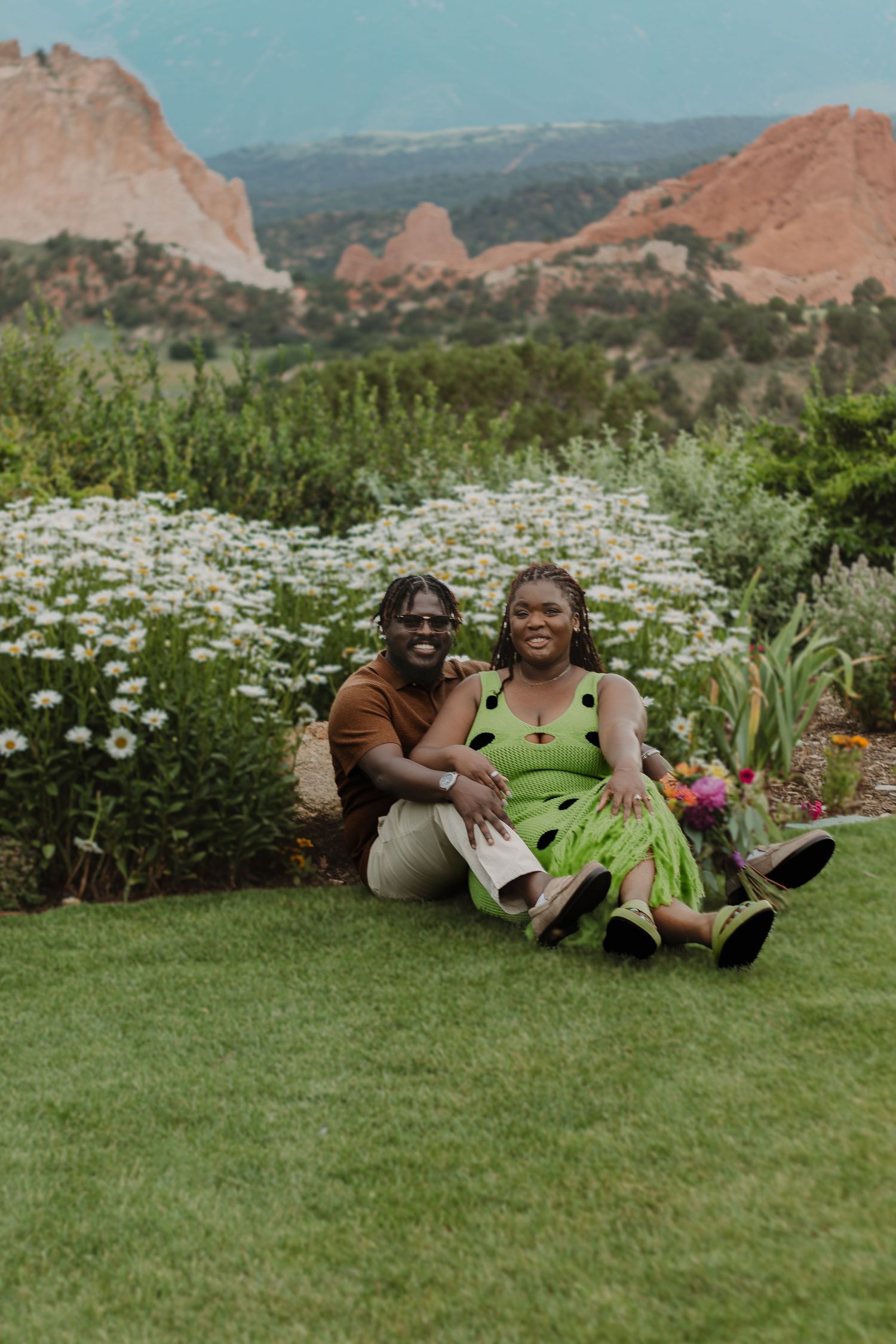 Two people lie in a meadow surrounded by wildflowers with majestic red rock formations visible in the distance.
