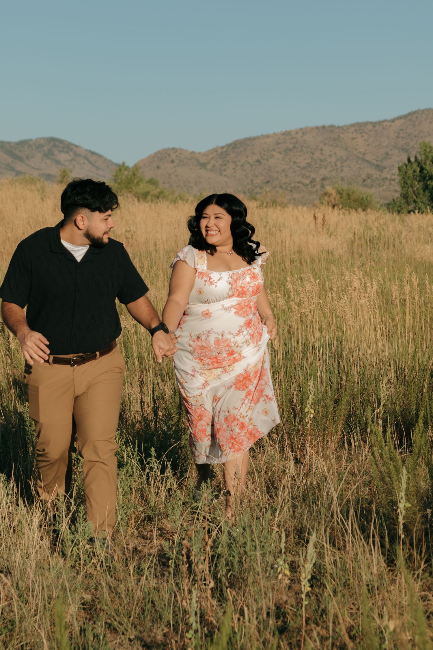 A couple walks together through tall grass with mountains visible in the desert landscape.