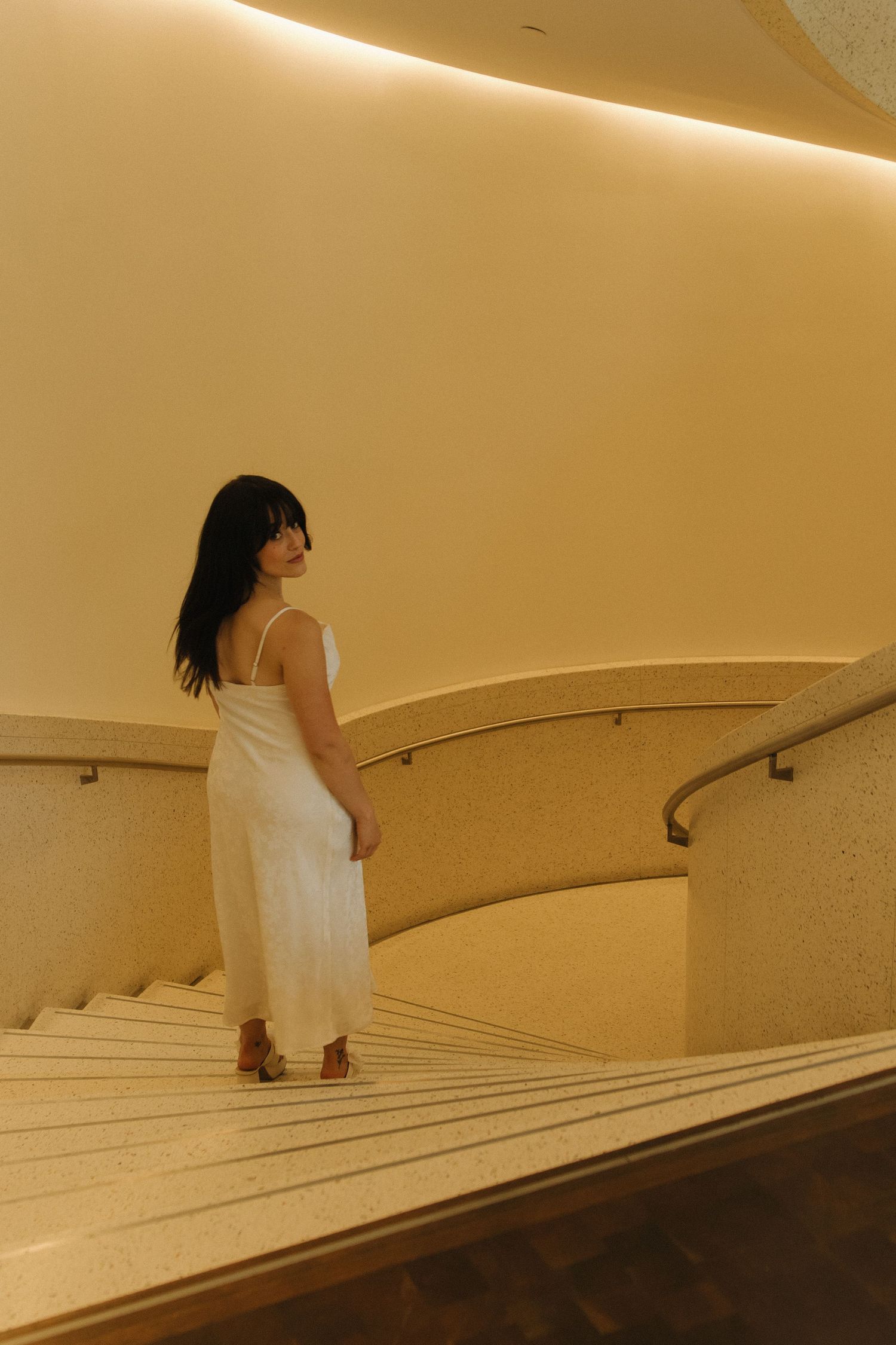 Figure in flowing white dress walking down curved marble staircase with handrail.