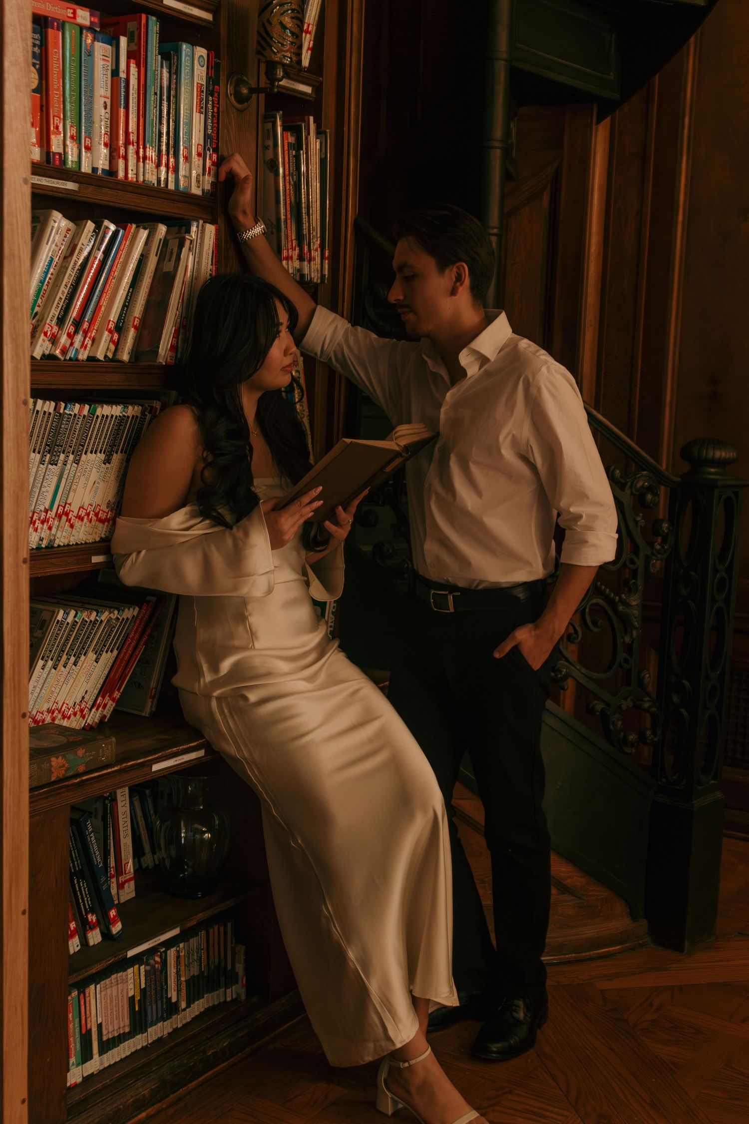 Couple in white outfits posing between library bookshelves in warm lighting.