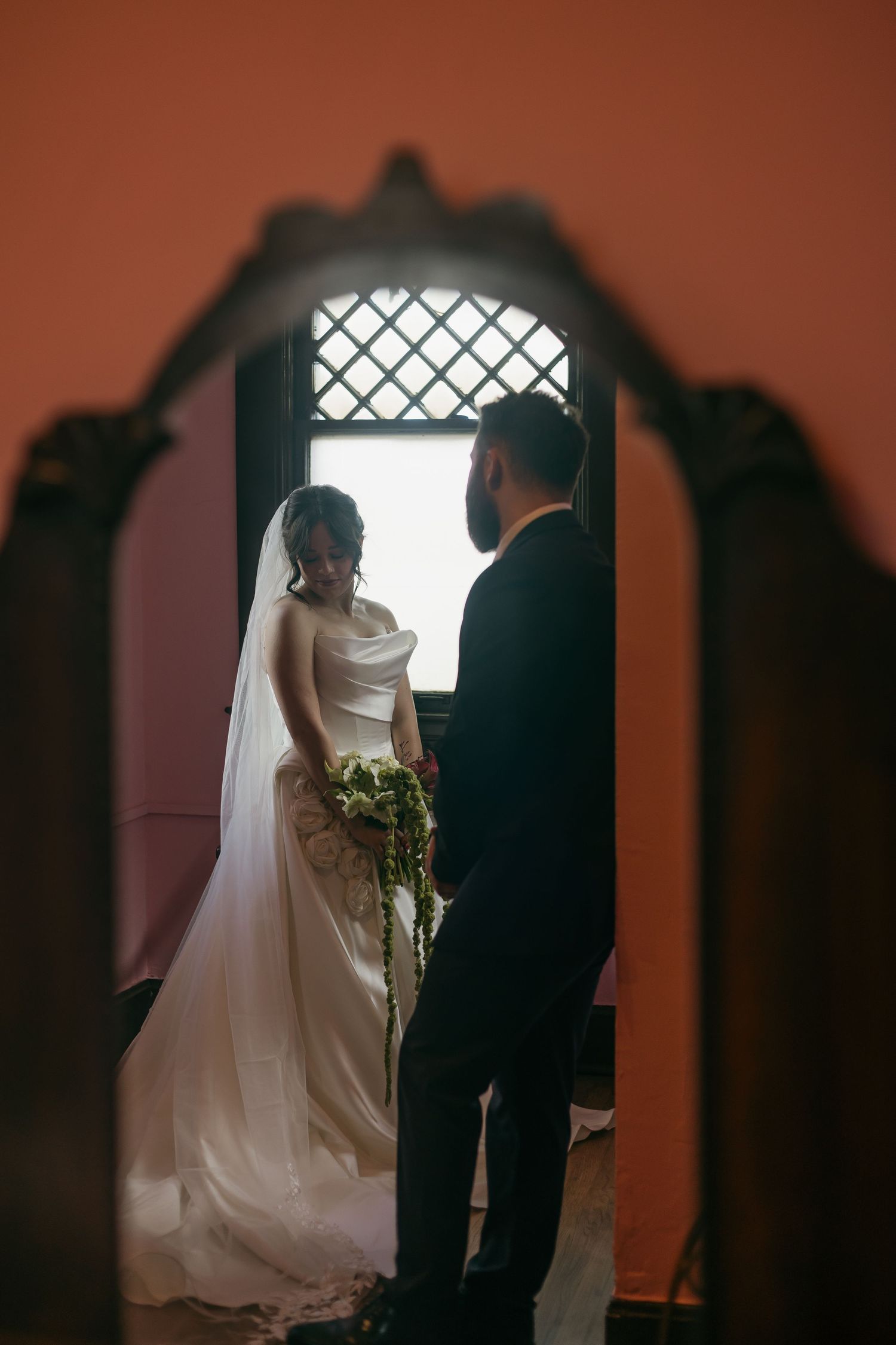 Silhouetted wedding couple stands before a window with cross pattern in warm-toned lighting.