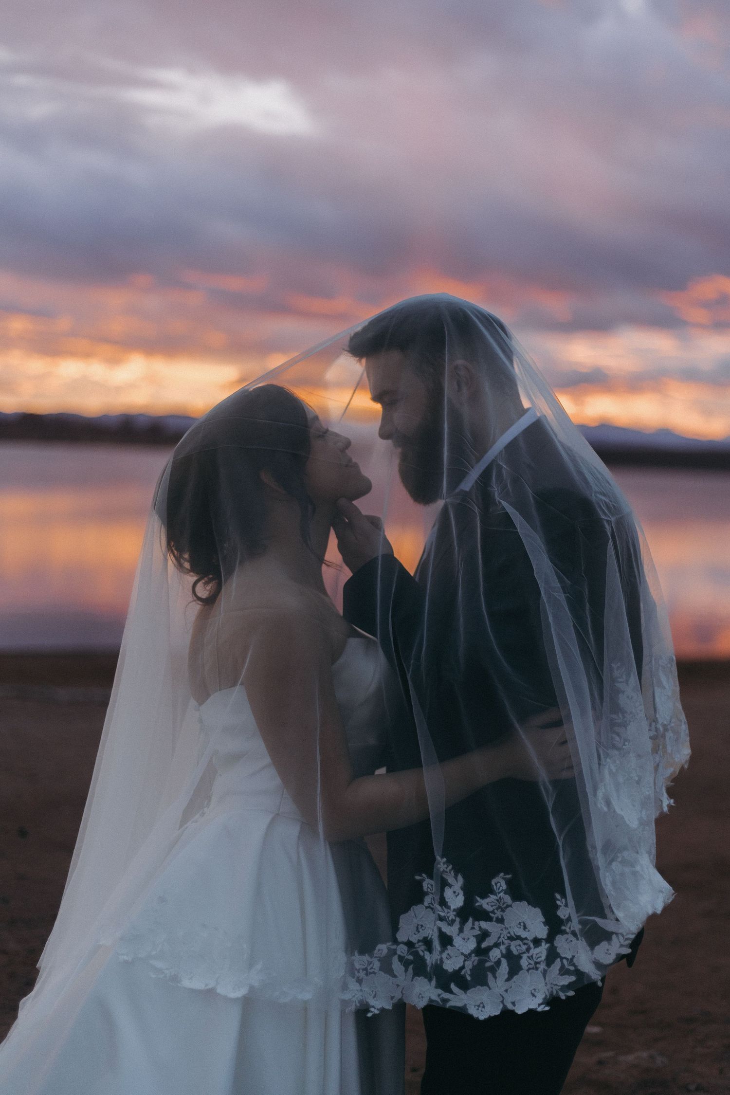 A couple in wedding attire shares an intimate moment at sunset by a lake.