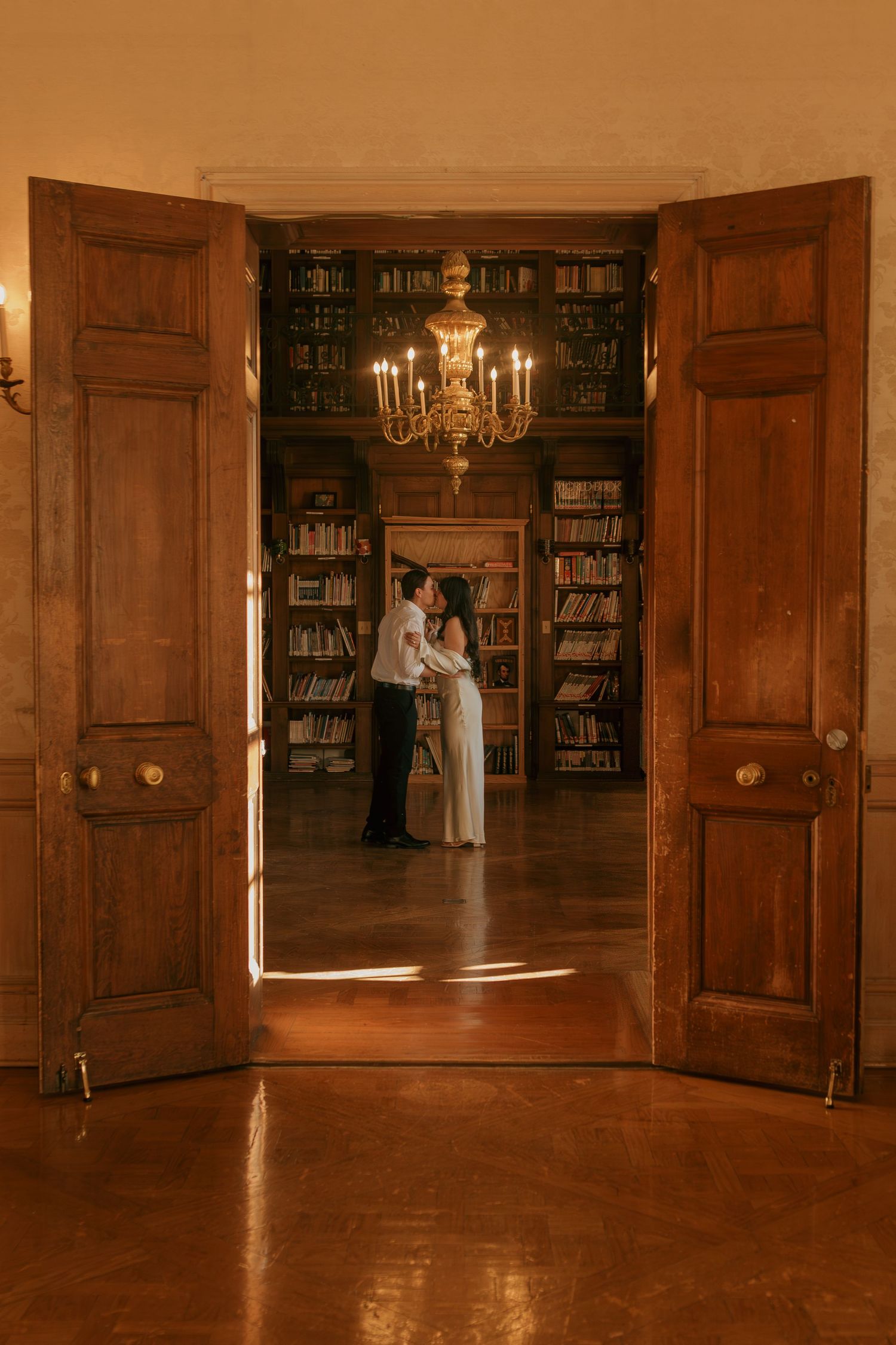 Wooden double doors open to reveal a grand library with chandelier and bookshelves.