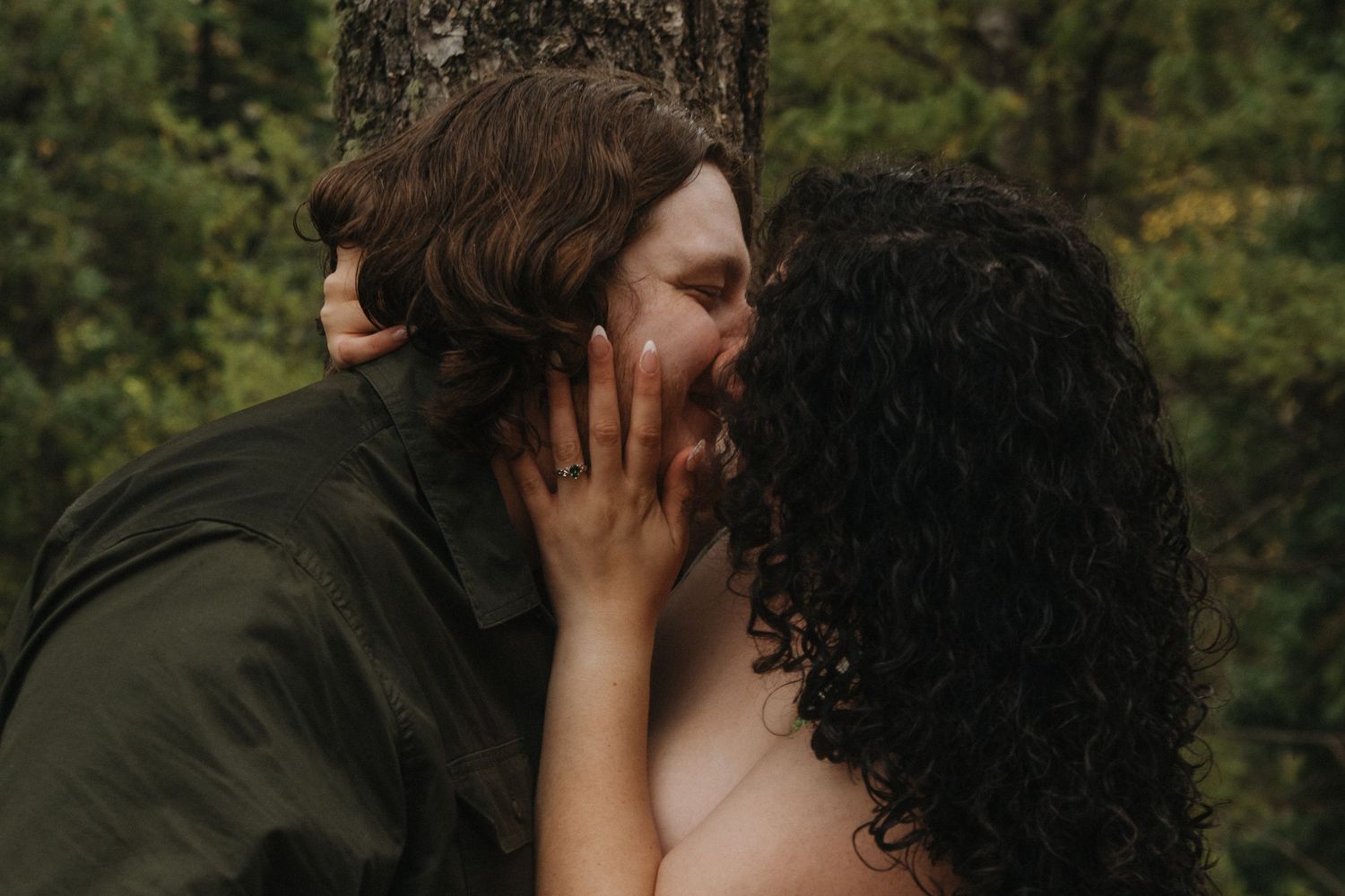 Two people share a romantic kiss while embracing in a lush forest setting at sunset.