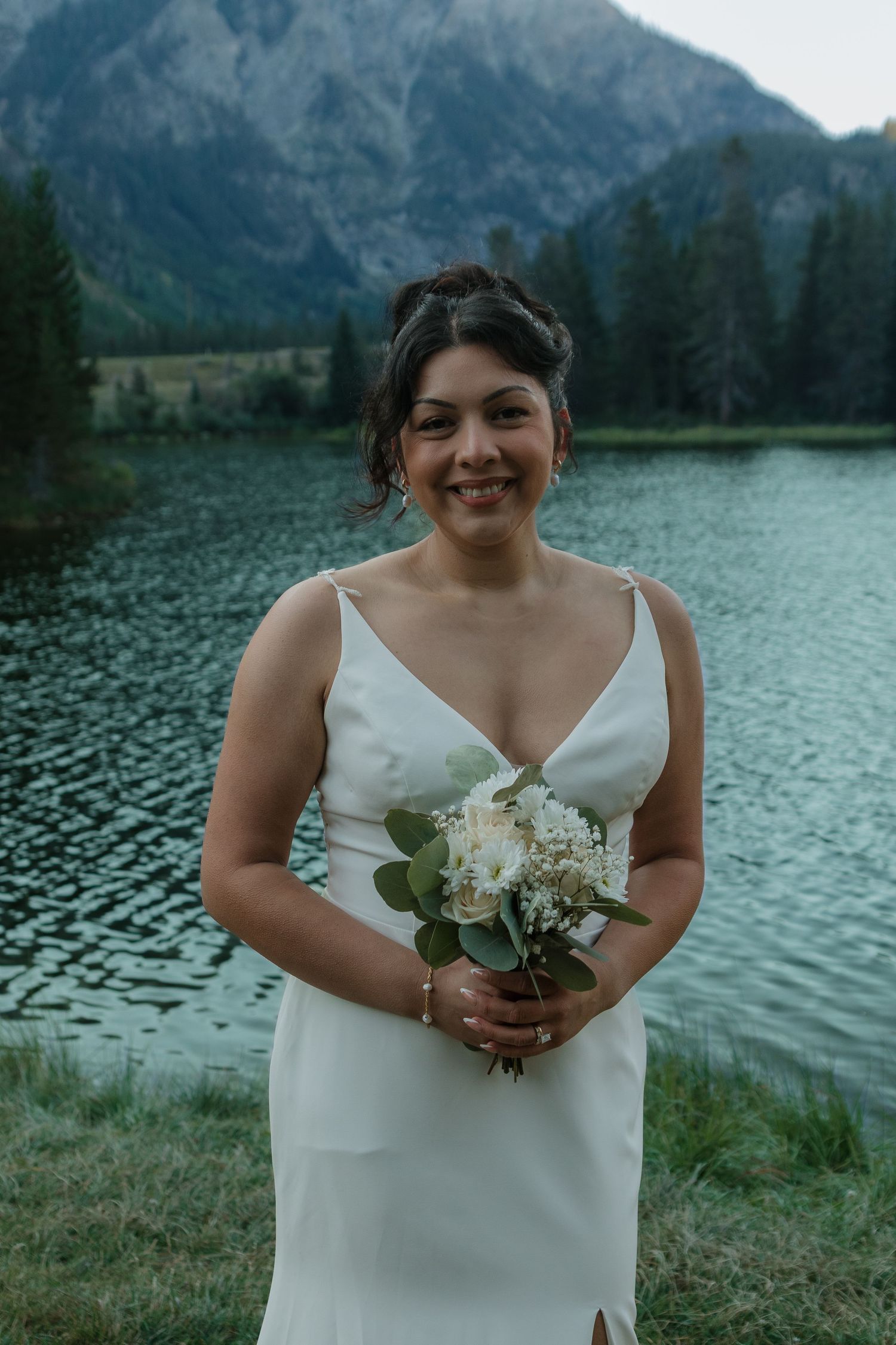 A person in a white wedding dress holds a small floral bouquet beside a mountain lake.