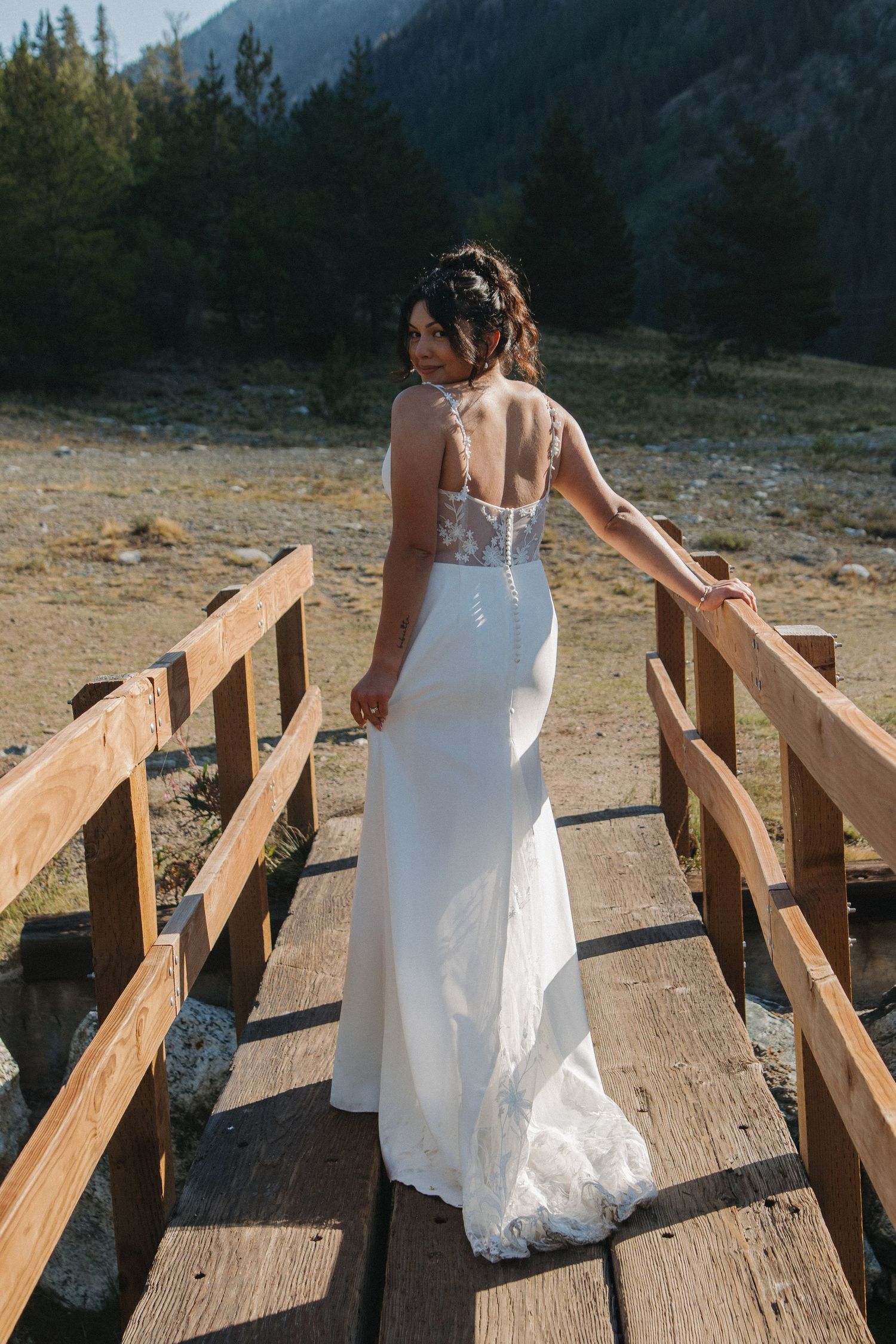 A white wedding dress flows gracefully across a rustic wooden bridge in a mountain setting.