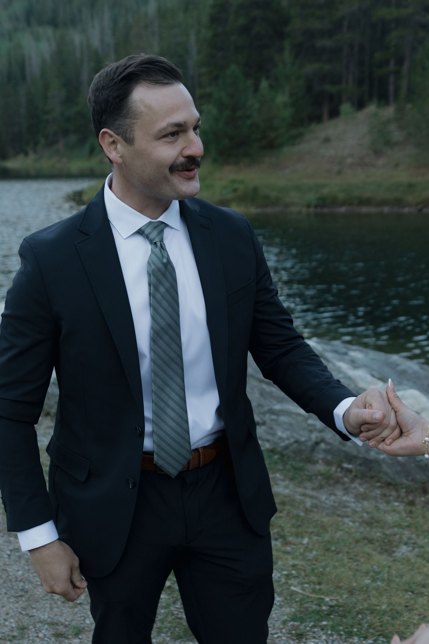 A person in a black suit jacket and tie gestures expressively against a scenic waterfront landscape.