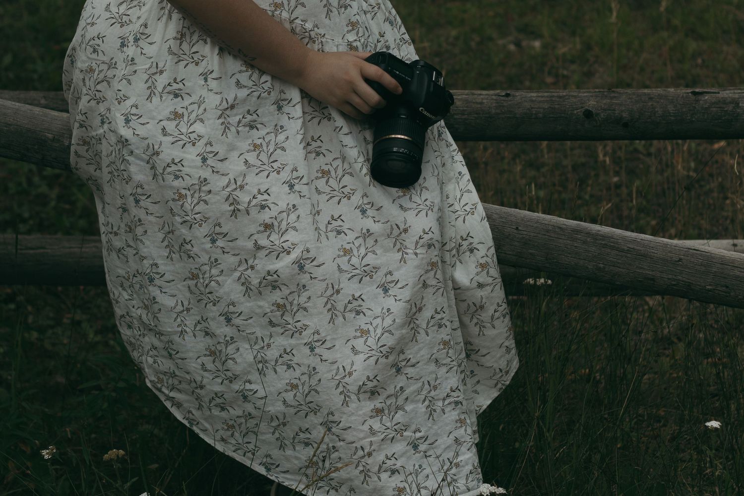A person in a floral dress stands in front of a rustic wooden fence in an outdoor setting.