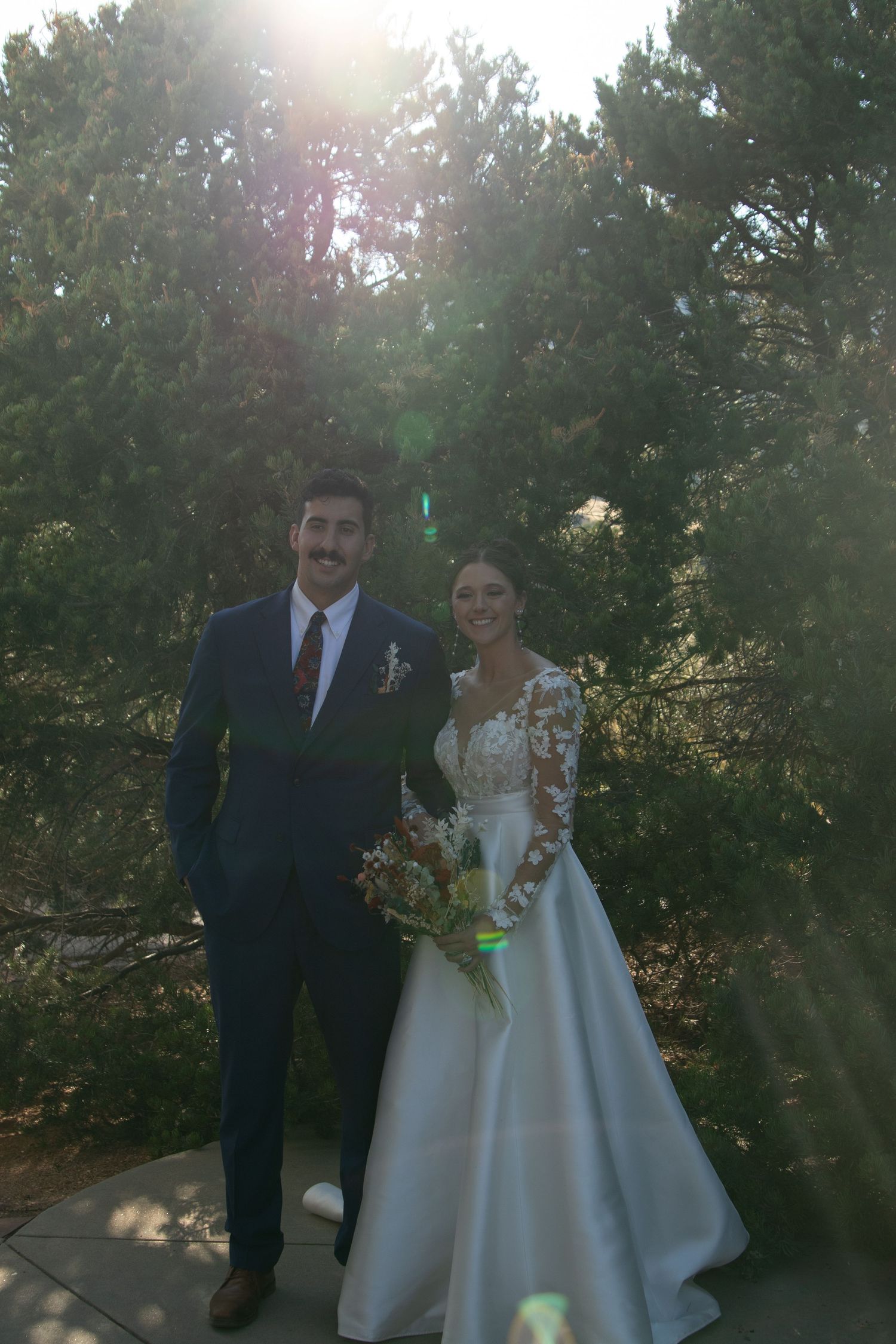 A wedding couple photographed against sunlit trees in the background.
