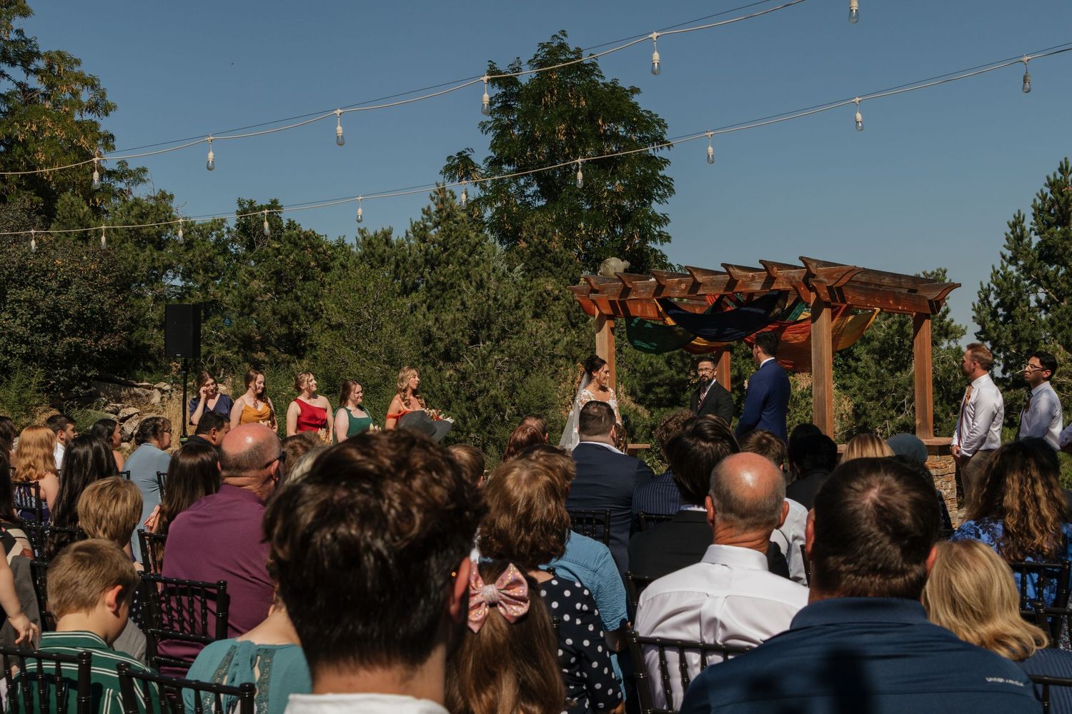 Outdoor wedding ceremony under blue skies with string lights and gathered guests.