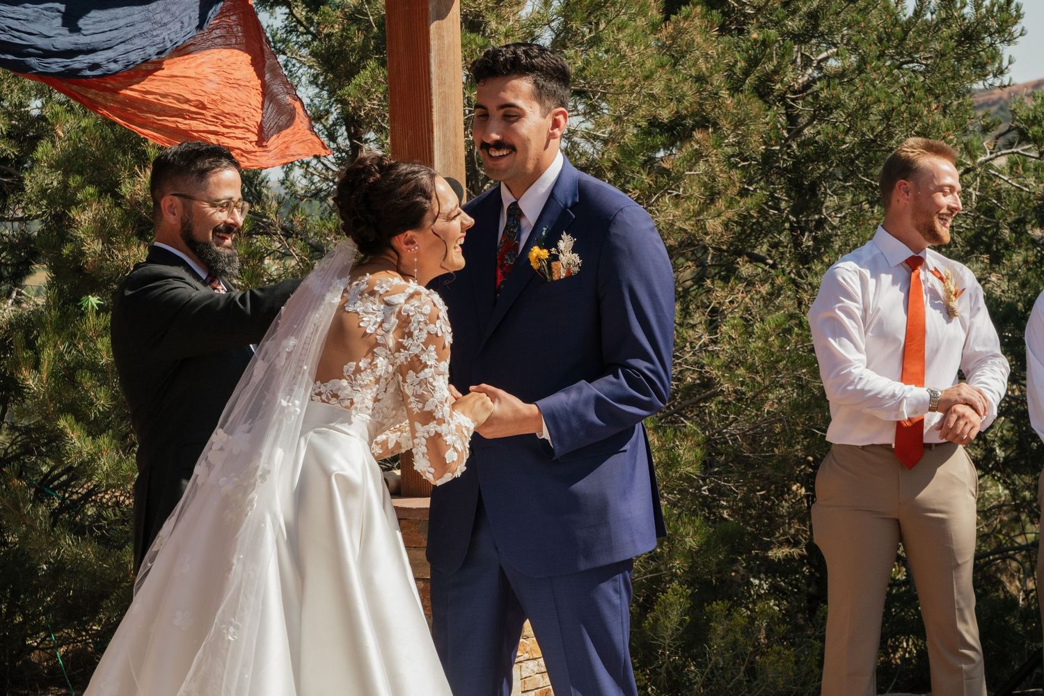 A wedding party stands outdoors by trees while posing for candid group photos.