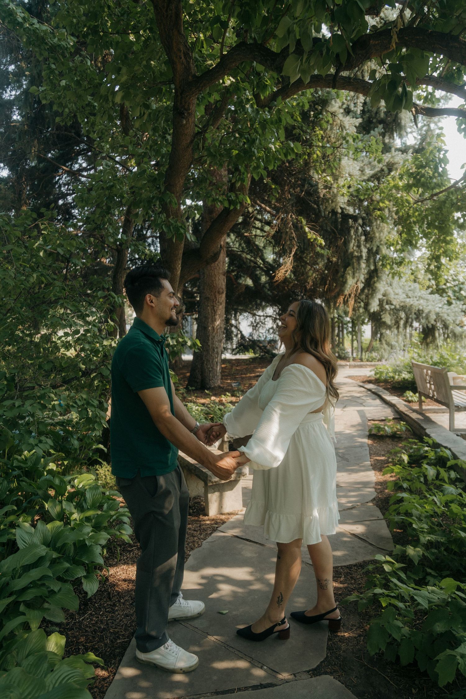 Couple holding hands and smiling on a garden path surrounded by lush green trees.