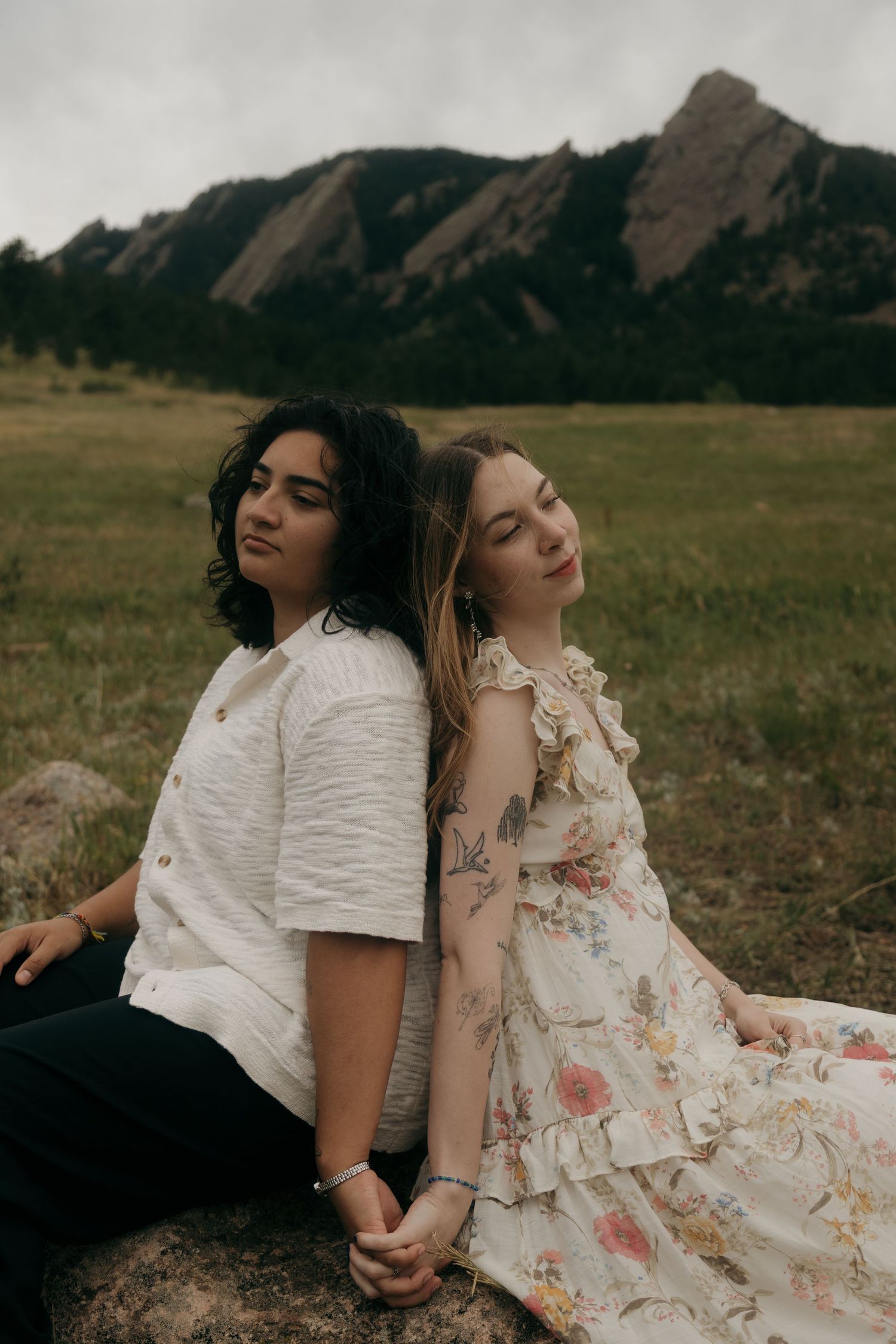 Two women sitting back-to-back on a rock in a grassy field with the Boulder Flatirons mountains behind them.